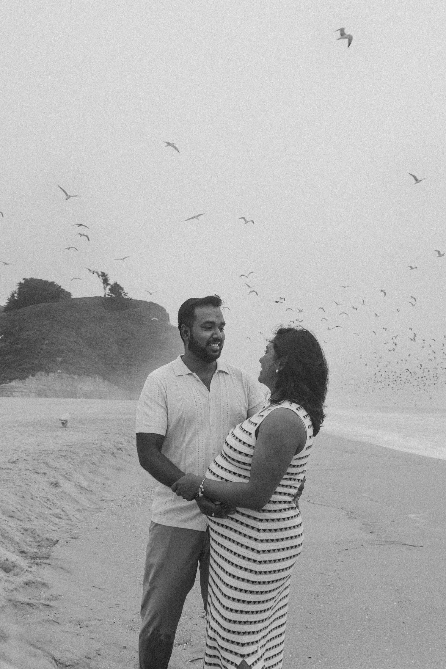 Couple embraces on beach with seagulls flying overhead against cloudy sky.