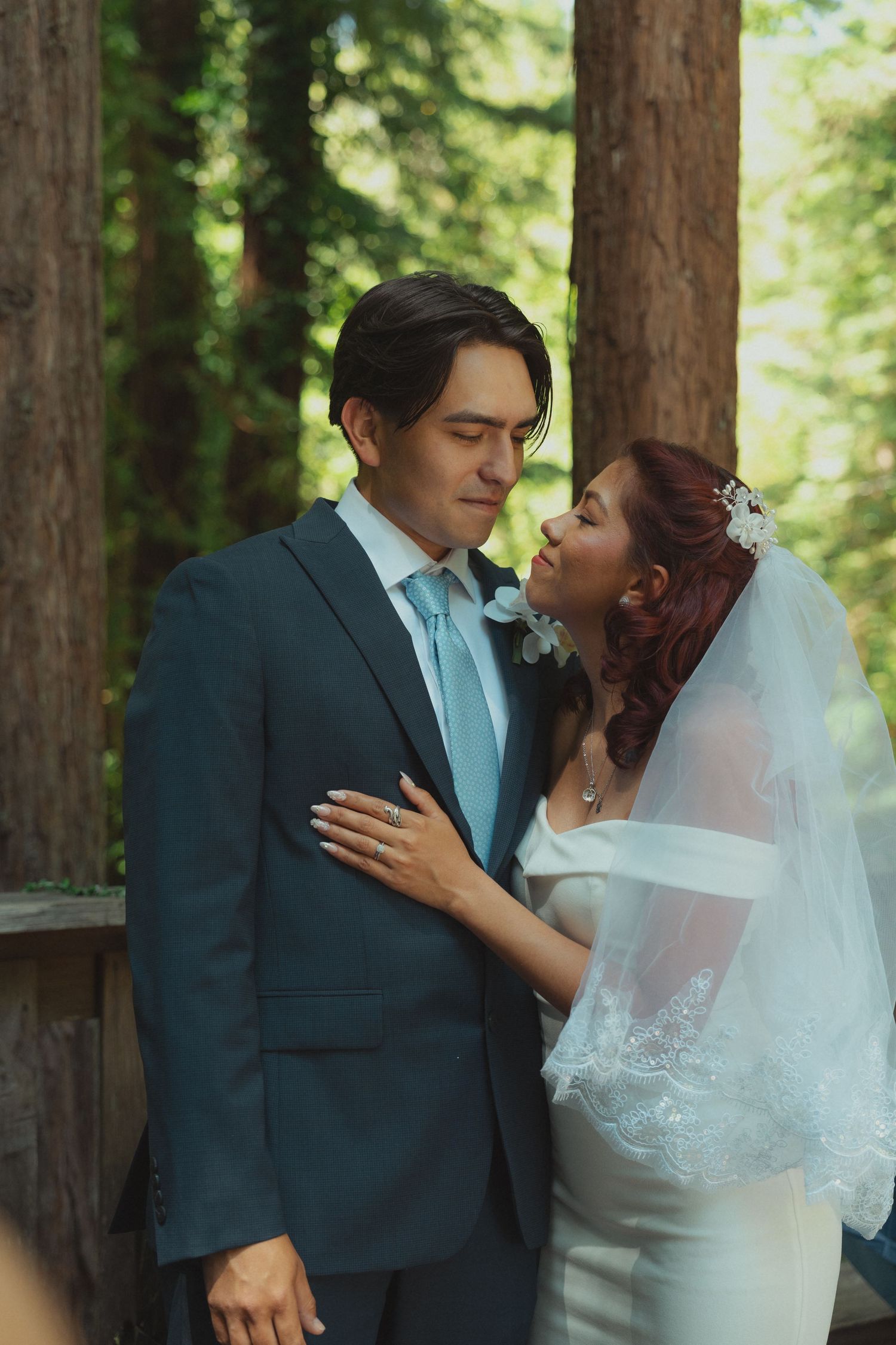 Bride and groom share an intimate moment among towering redwood trees, she in a white gown with veil, he in a blue suit.