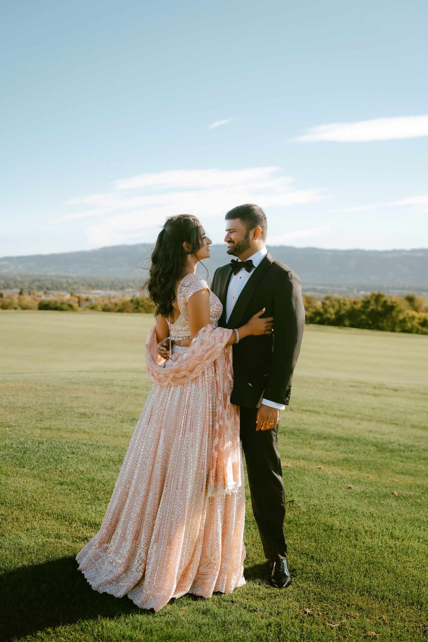 Couple in elegant wedding attire standing on green lawn with mountain backdrop on sunny day.