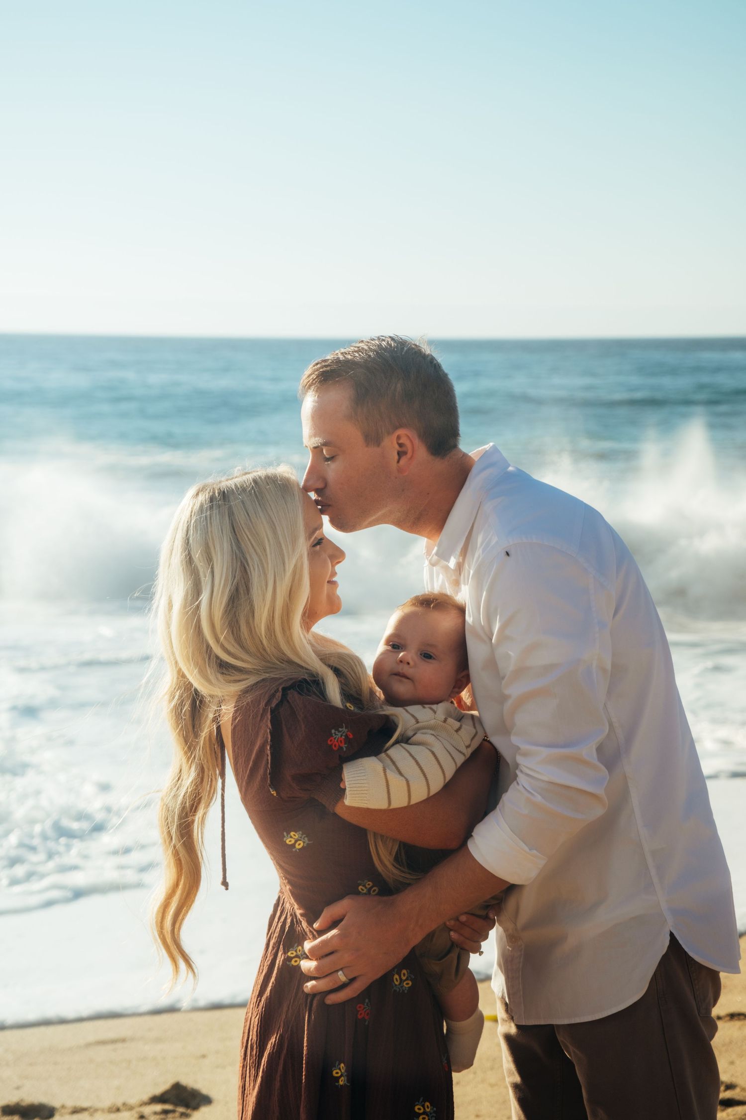 Loving family embraces while standing on sandy beach with ocean waves in background.