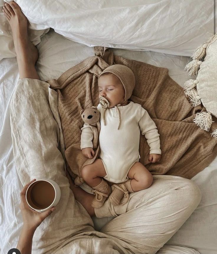 A baby in neutral-toned clothing lies on a beige blanket with a coffee cup placed nearby.