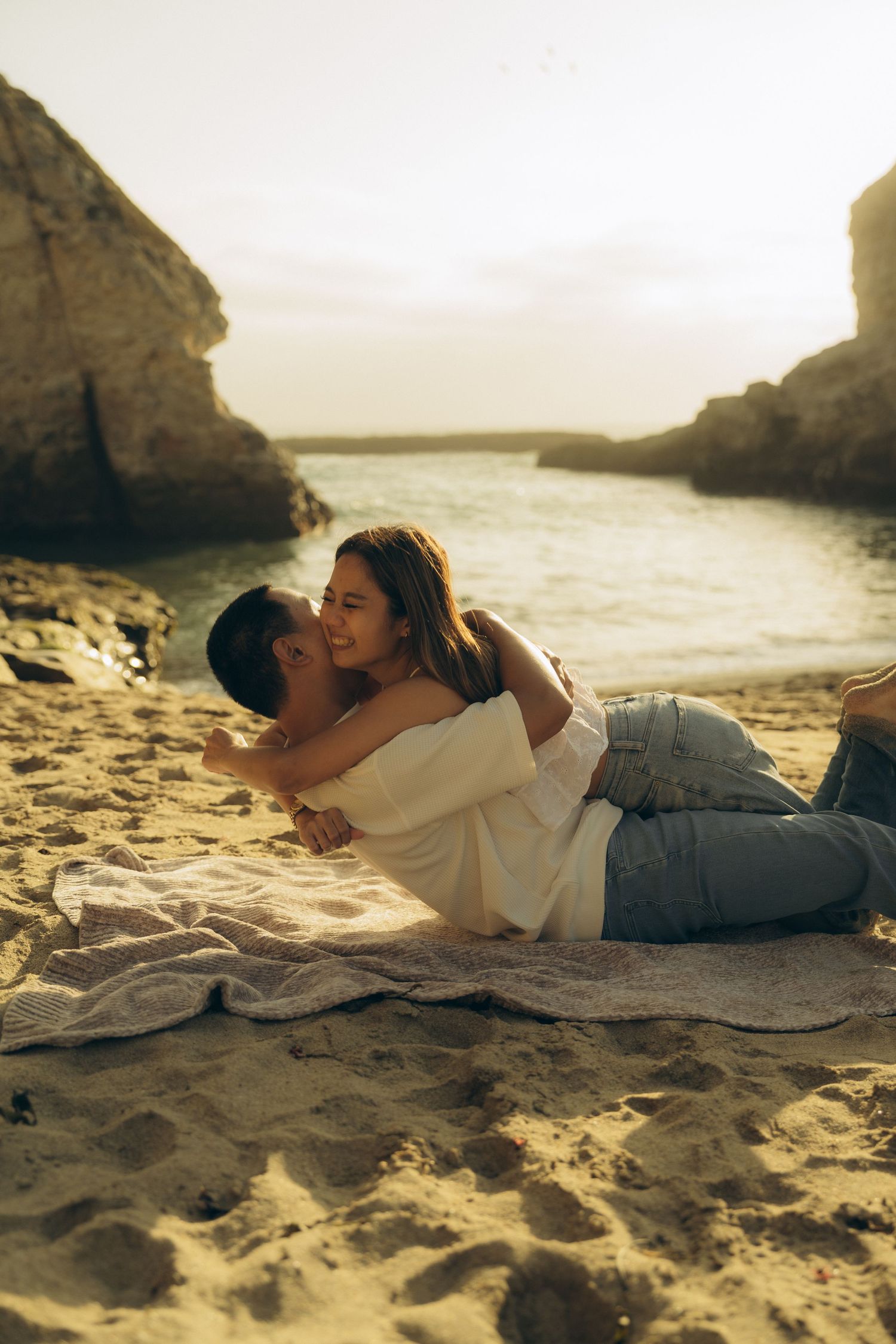 Romantic couple embracing on sandy beach at sunset, lying on blanket near rocky cove with turquoise water.