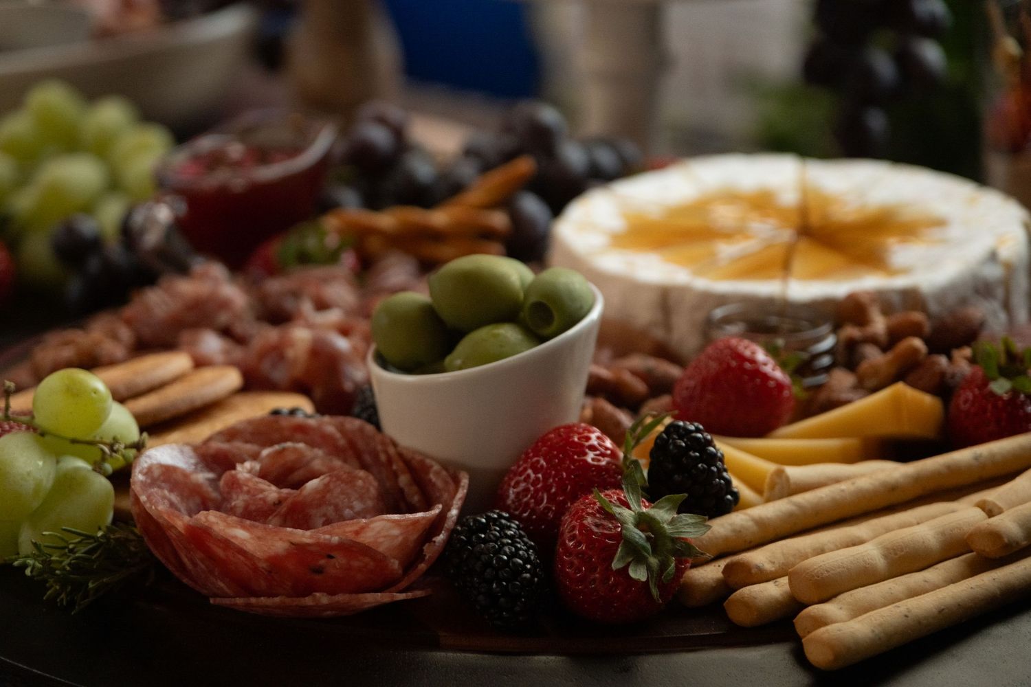 Charcuterie board with meats, cheeses, fruits and breadsticks arranged on dark surface.