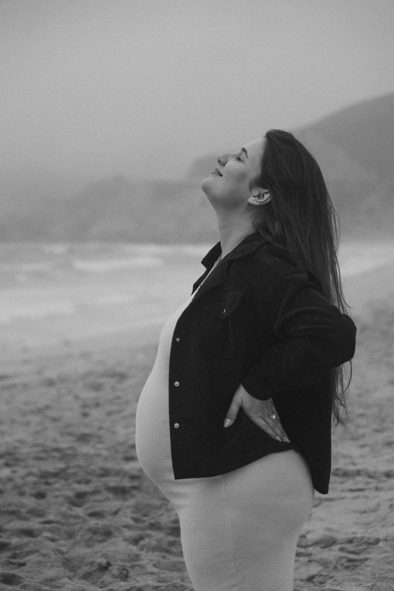 A pregnant woman in a white dress poses expressively on a beach during a photoshoot.