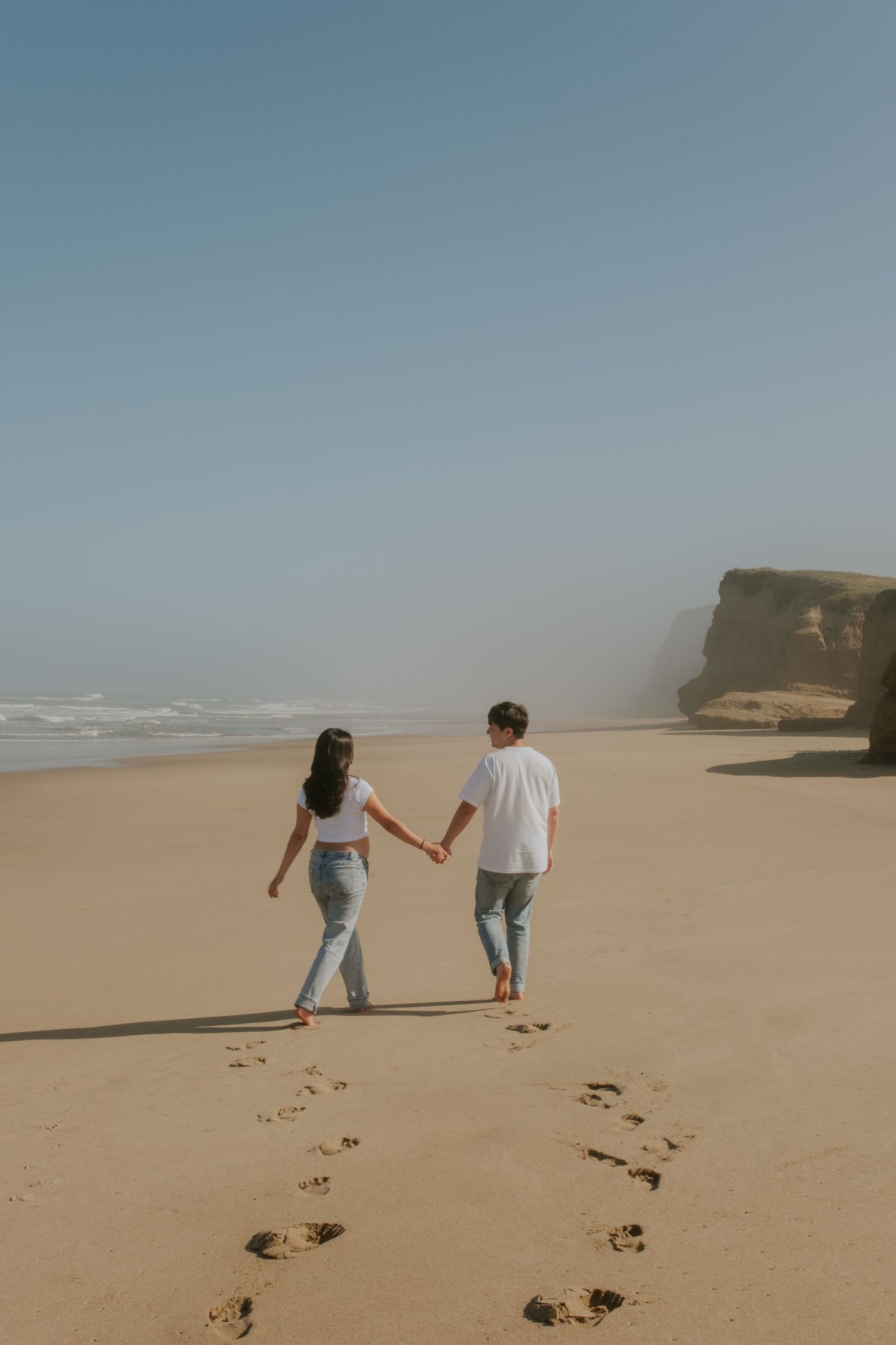 Footprints trail behind a couple walking hand in hand on a vast sandy beach.