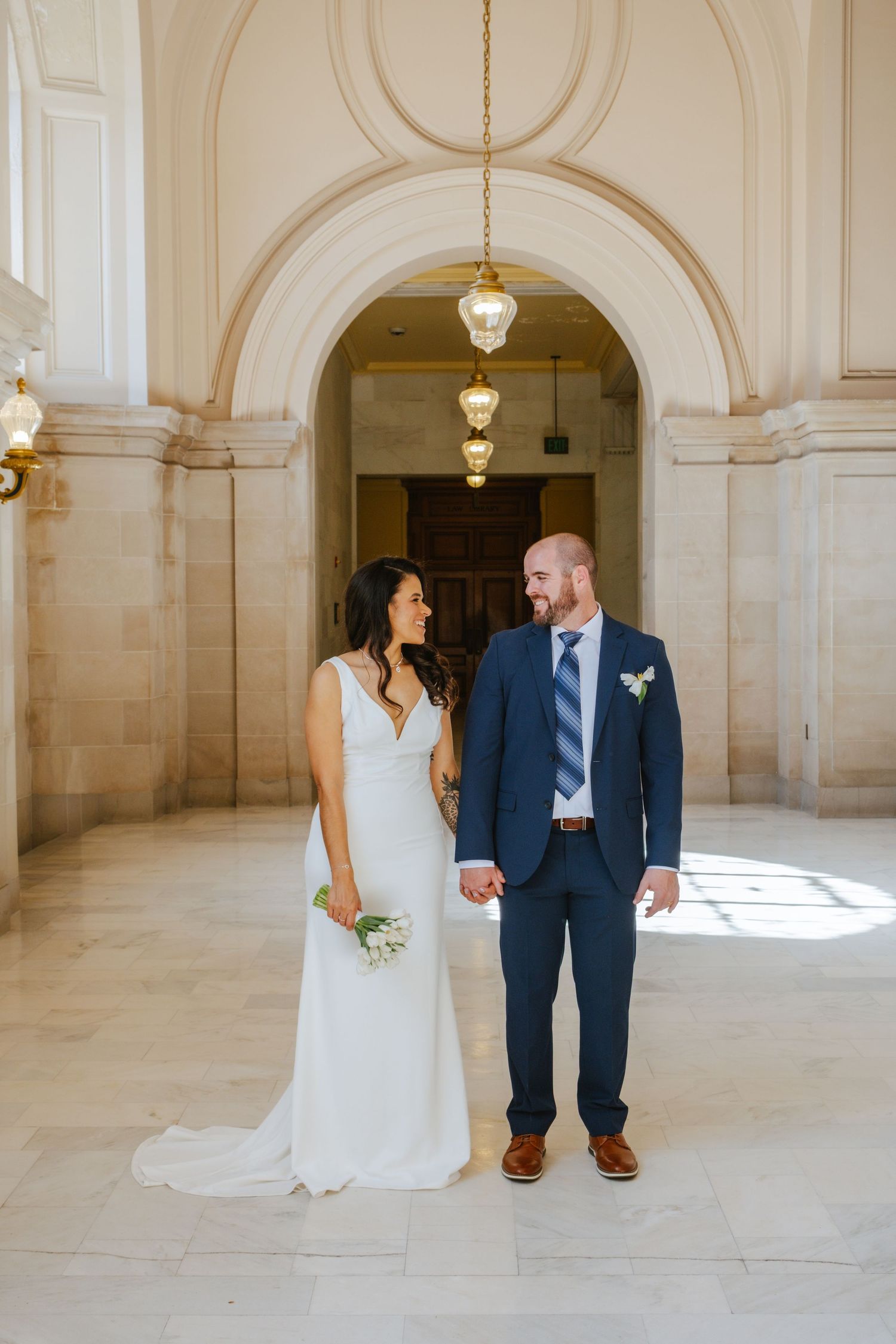 Newlywed couple poses for romantic portraits in an elegant white marble hallway with hanging pendant lights.