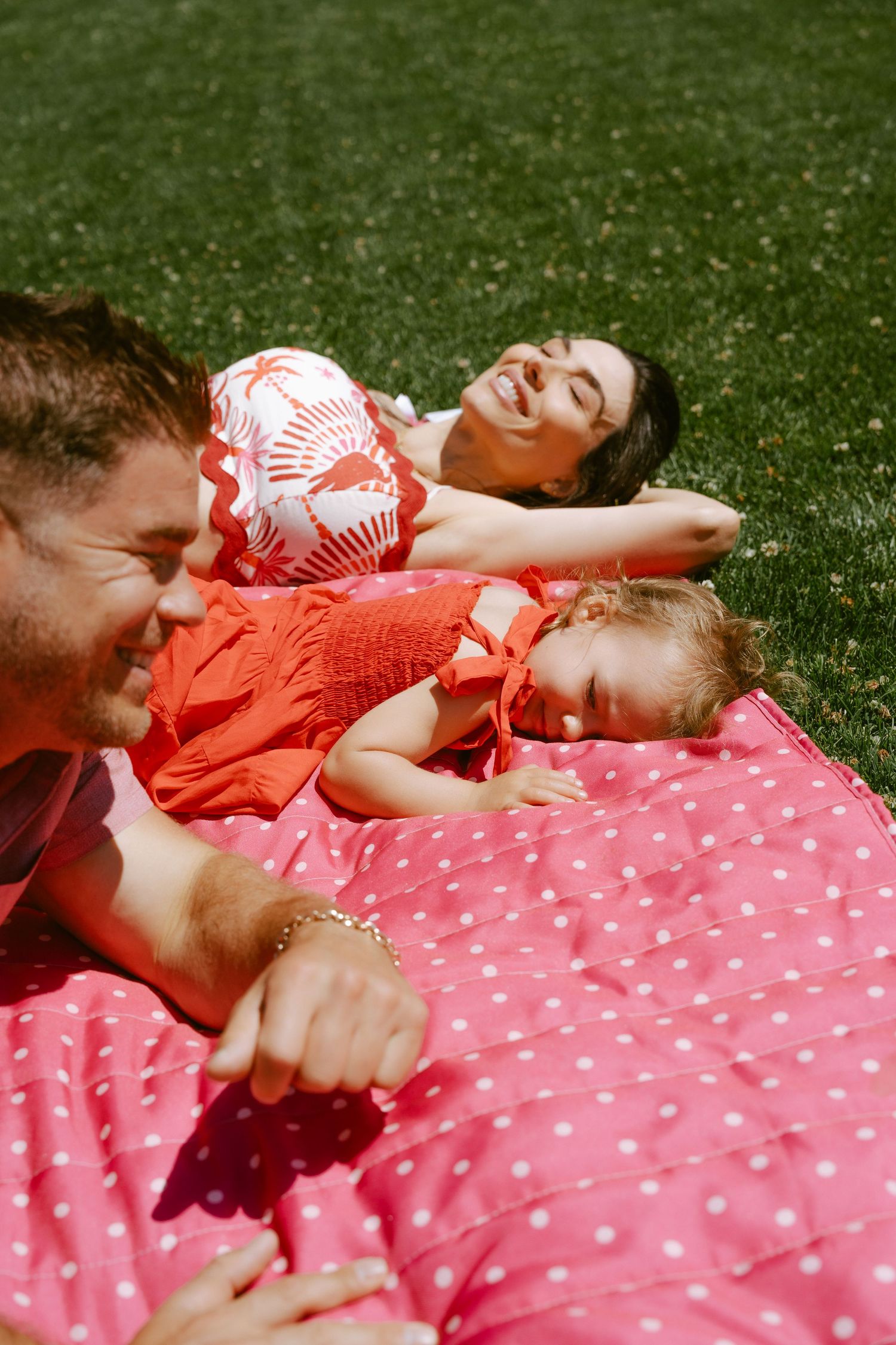 Family relaxing on a pink polka dot blanket in sunny grass, smiling parents with toddler in matching red outfits.