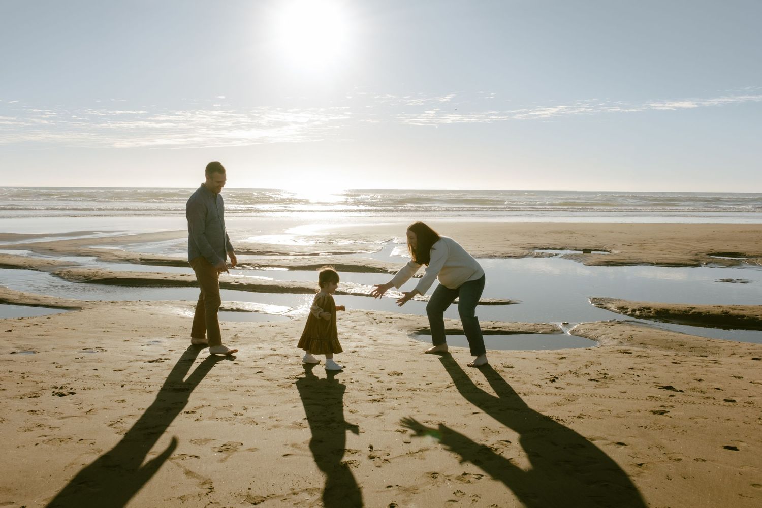 Family of three enjoying a sunny beach day, with parents playing with toddler near tidal pools at sunset.