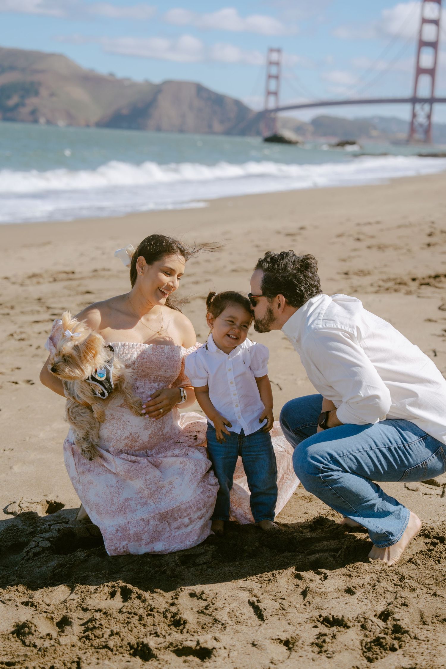Family maternity photo on Baker Beach with Golden Gate Bridge, parents and toddler with small dog on sunny day.