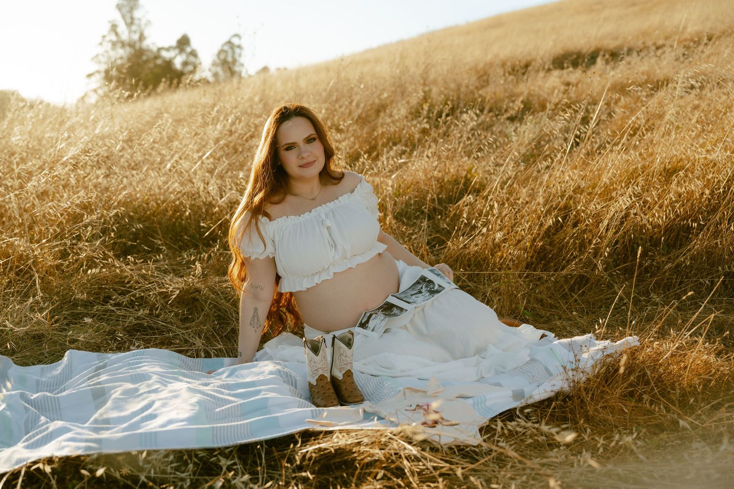 Pregnant woman in white outfit sitting on blanket in golden grass field during sunny maternity photo session.
