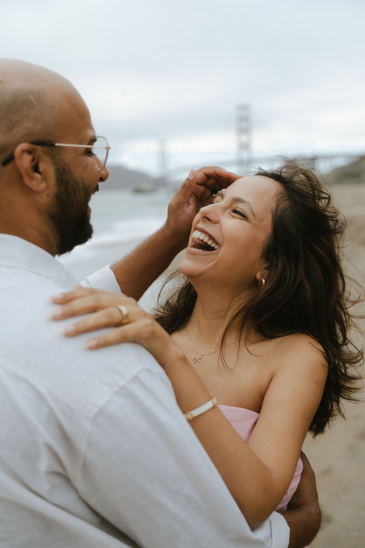 Couple laughing together on Baker Beach with Golden Gate Bridge in background during engagement photo session.