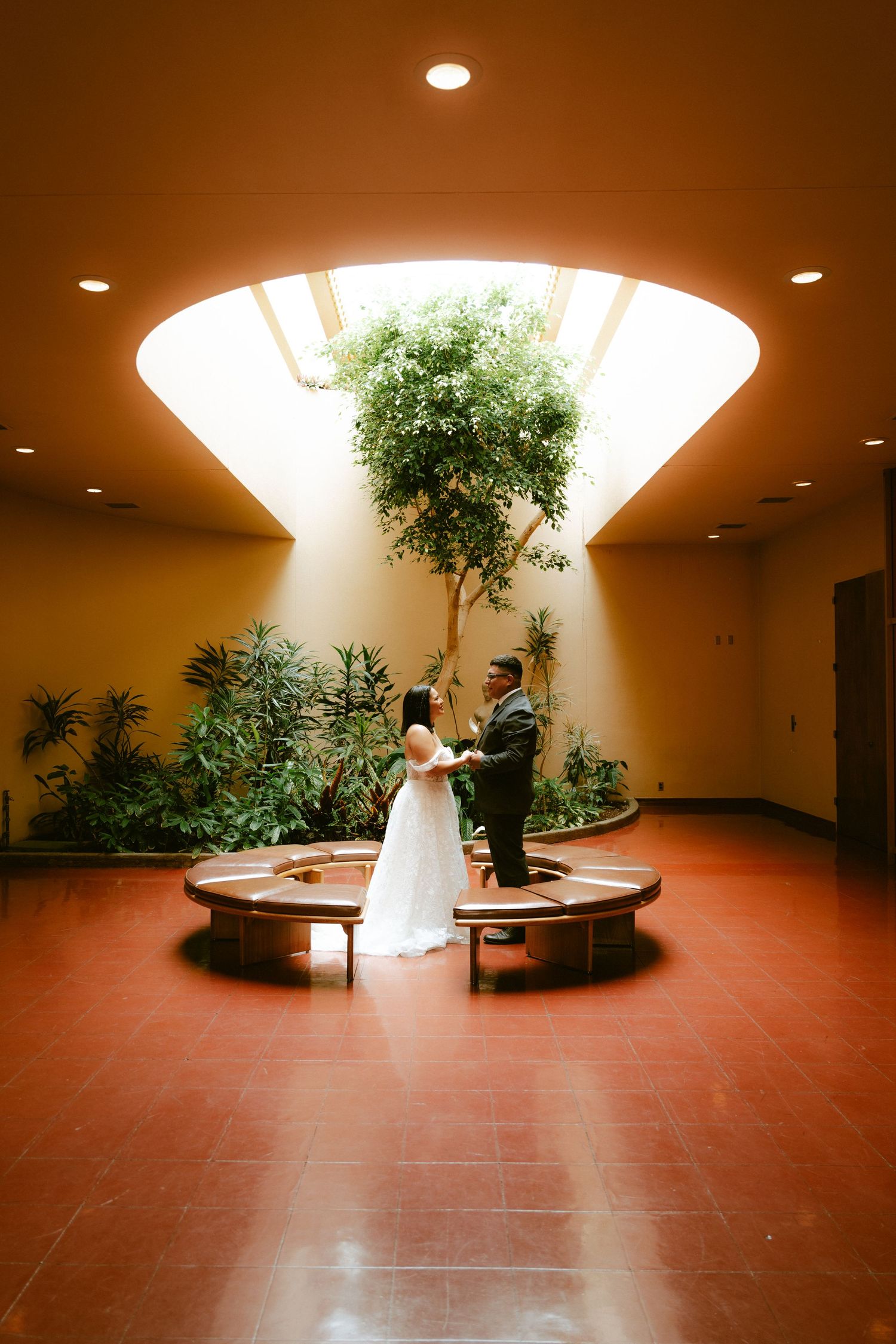 Bride and groom exchange vows in elegant indoor atrium with lush trees and warm lighting during intimate wedding ceremony.