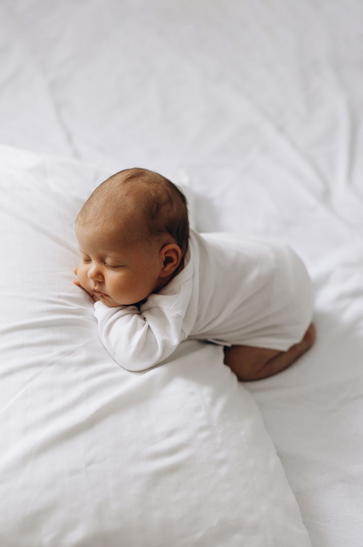 A peaceful sleeping newborn baby in white clothing rests on crisp white bedding.
