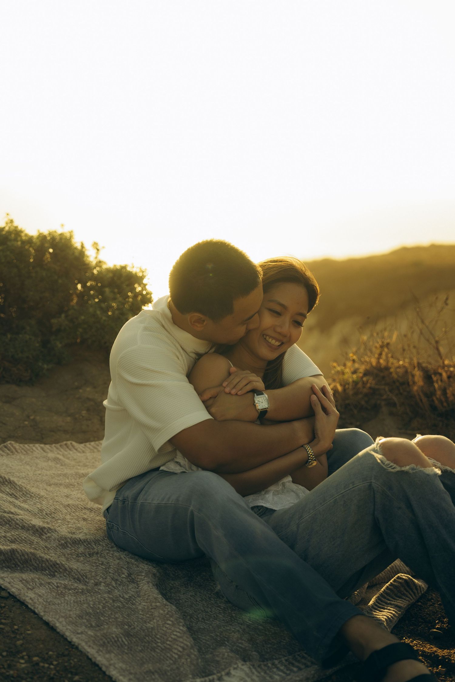 Couple sharing a romantic embrace at golden hour sunset on a blanket outdoors.