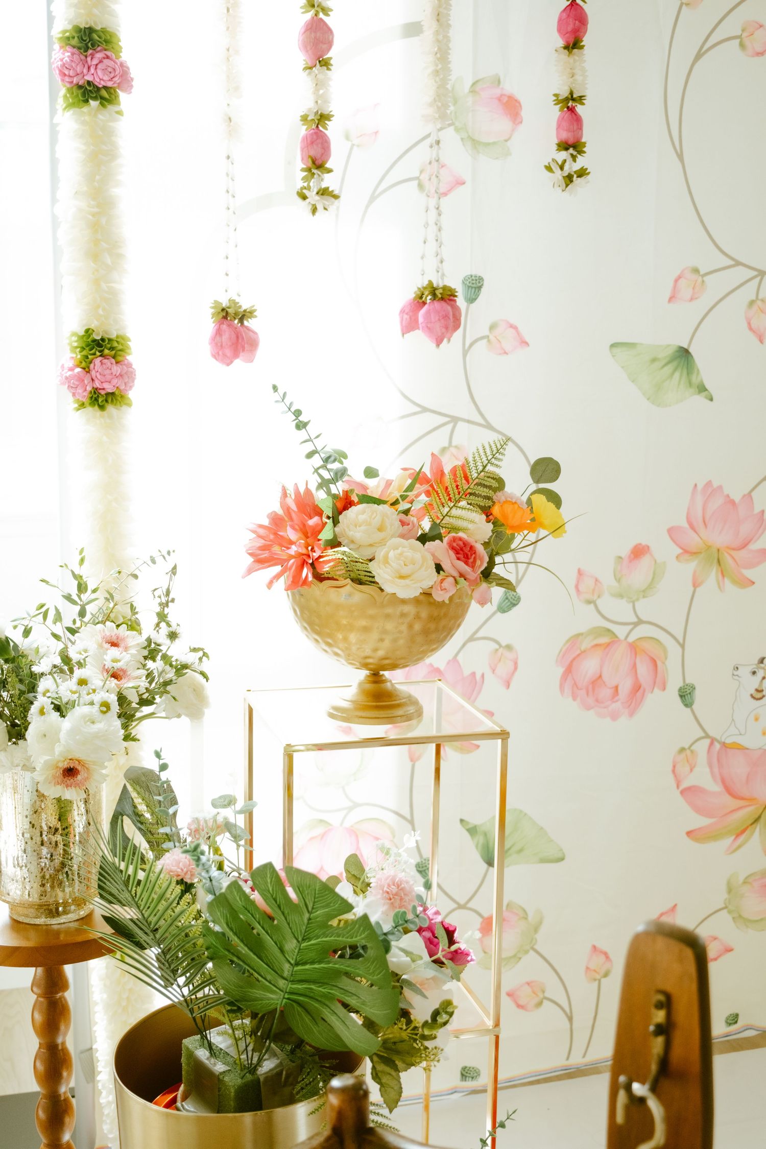 Gold vase with pink and white floral arrangement on stand, flower garlands, and botanical wallpaper in elegant decor.