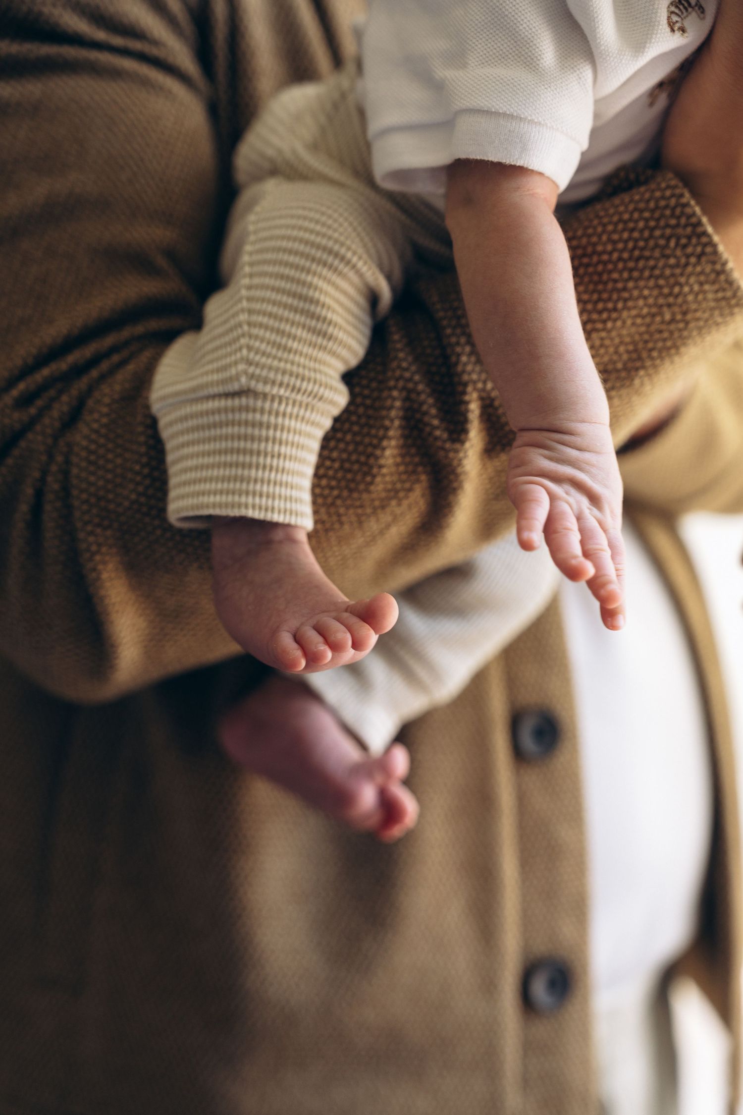 Close-up of a newborn baby's tiny feet and hands resting on a parent's arm in a brown knit cardigan.