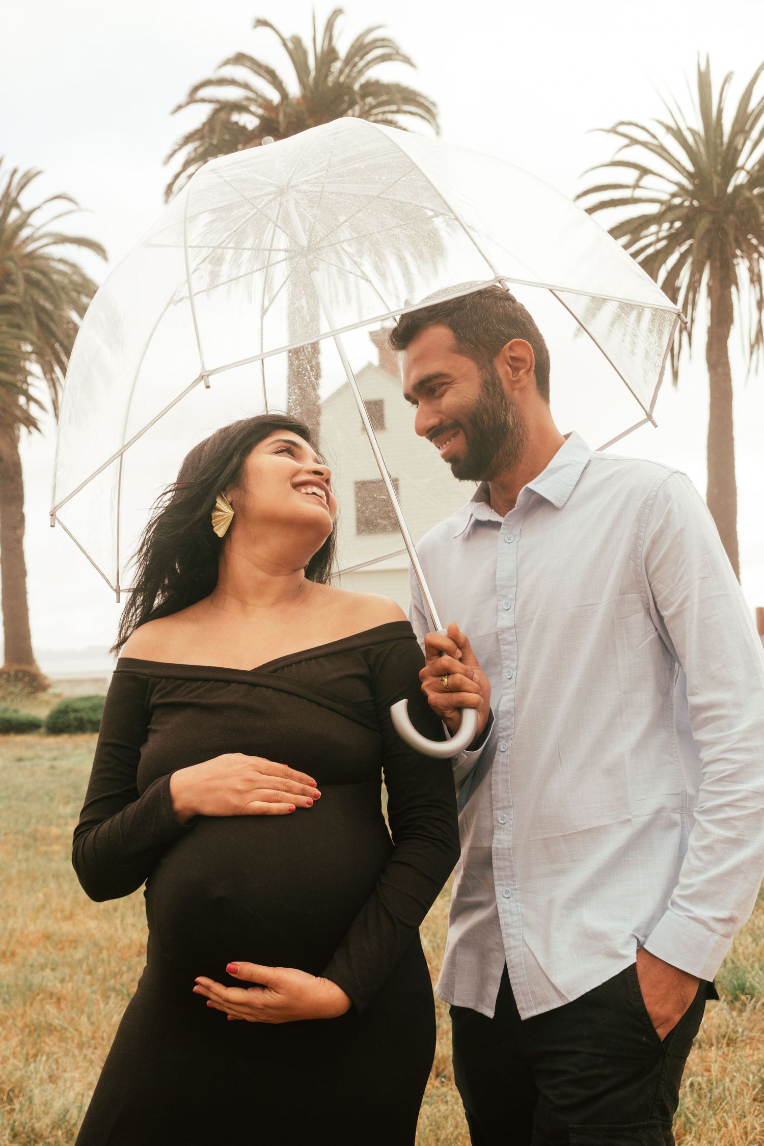 Couple with clear umbrella posing near palm trees on misty day.