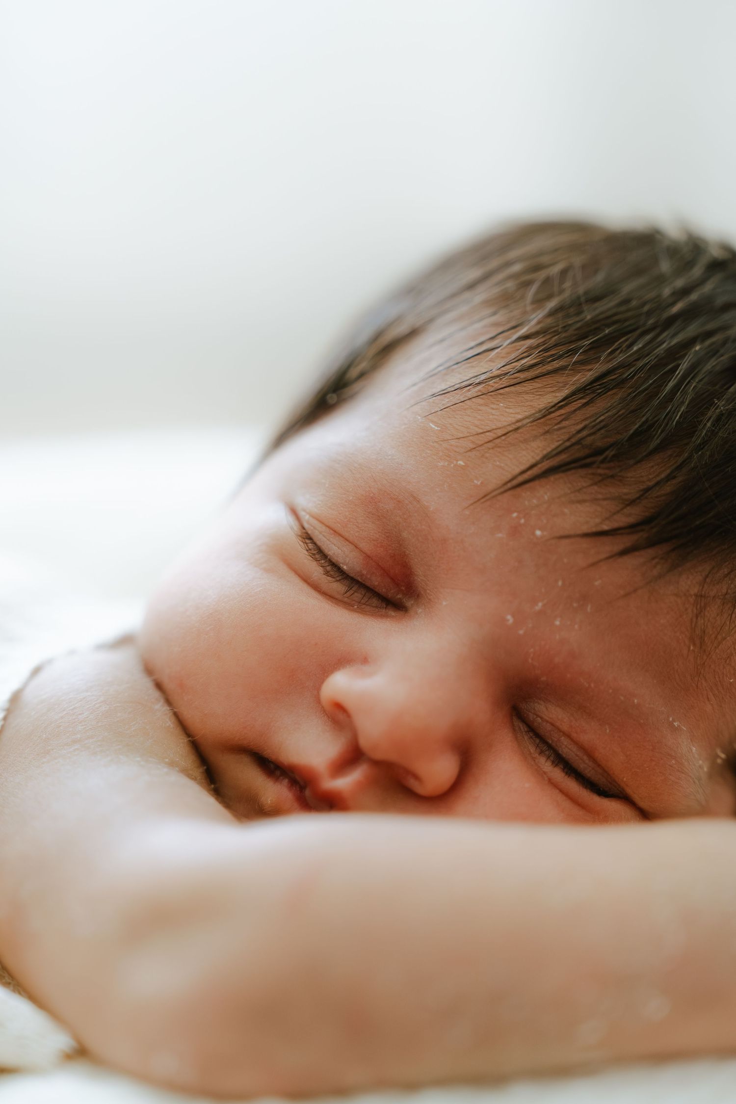 Peaceful newborn baby sleeping with dark hair, eyes closed, resting on hands on a soft white surface.