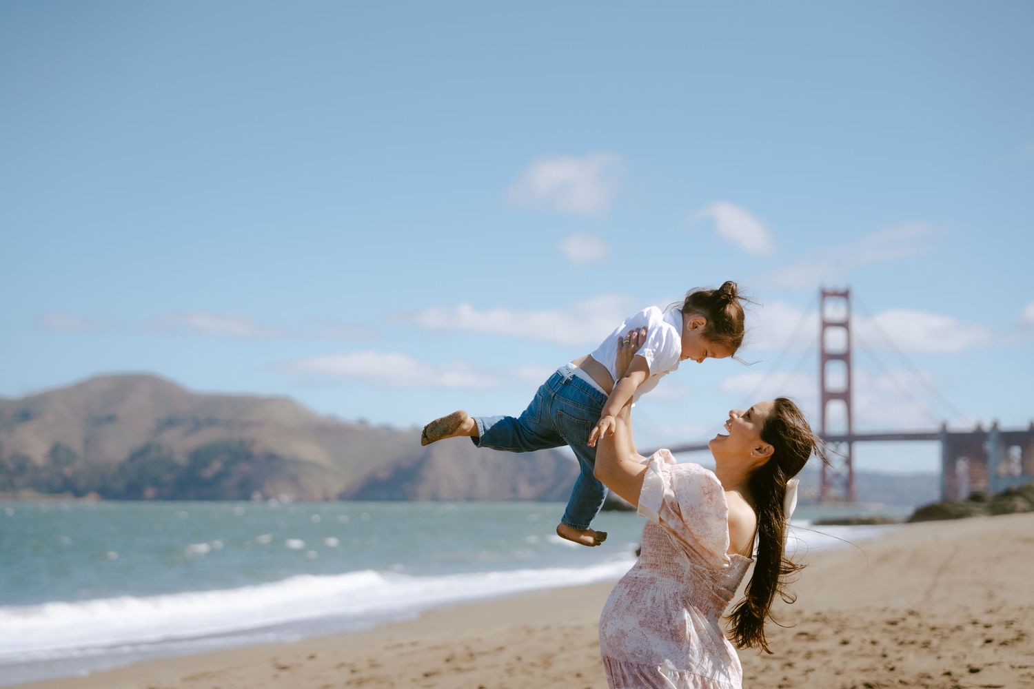 Mother lifting toddler on Baker Beach with Golden Gate Bridge backdrop on sunny day.