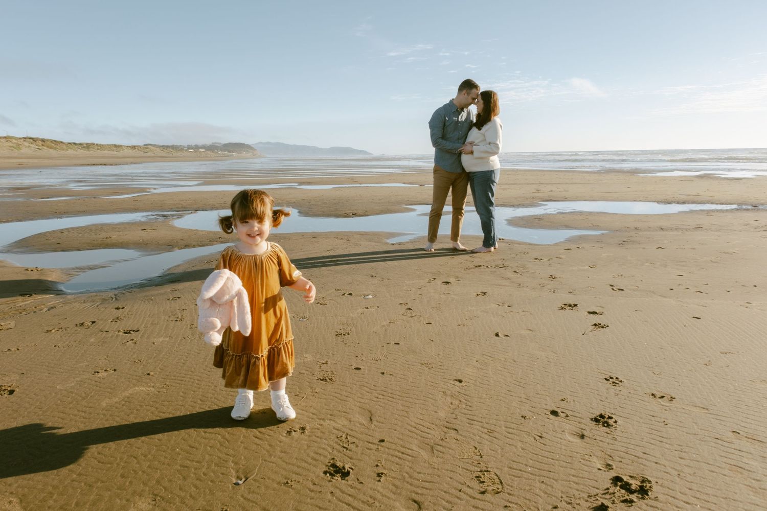 A toddler girl in a rust dress walks on a sandy beach while her parents embrace in the background.