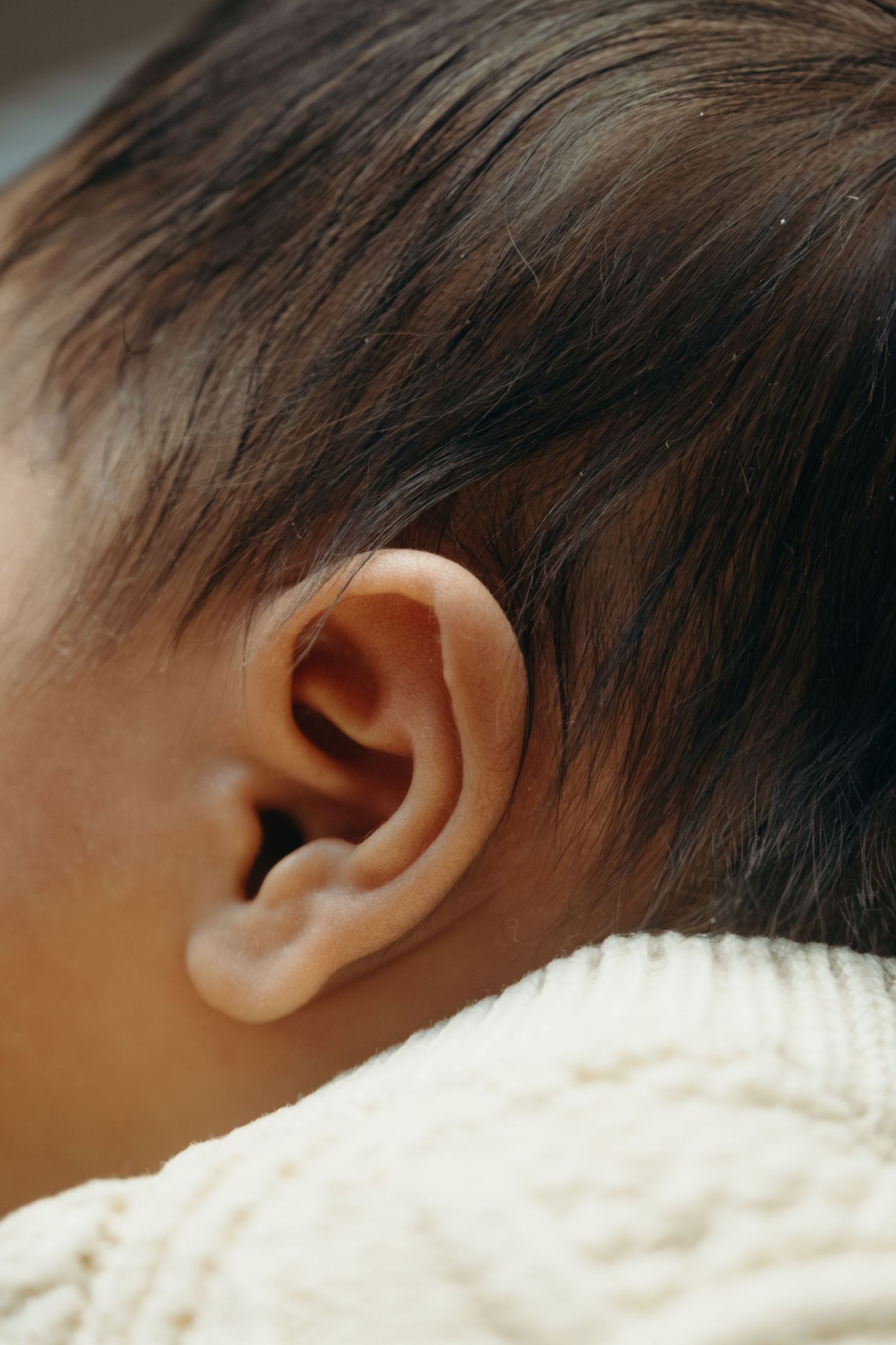 Close-up of a baby's ear with dark hair, wearing a cream knit sweater.