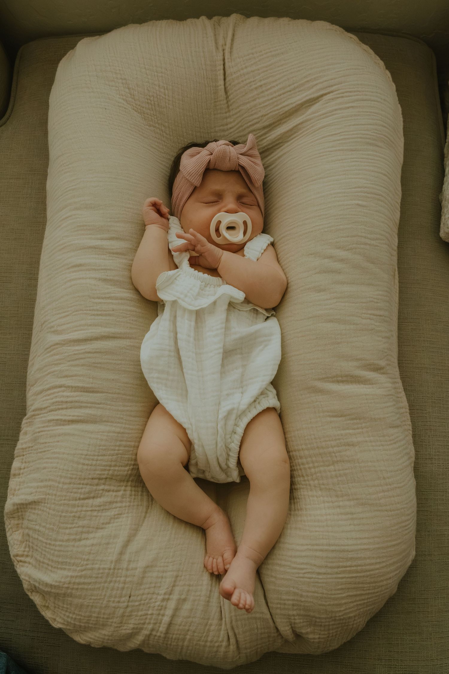 Sleeping newborn baby girl in white onesie and pink bow headband with pacifier on beige nursing pillow.