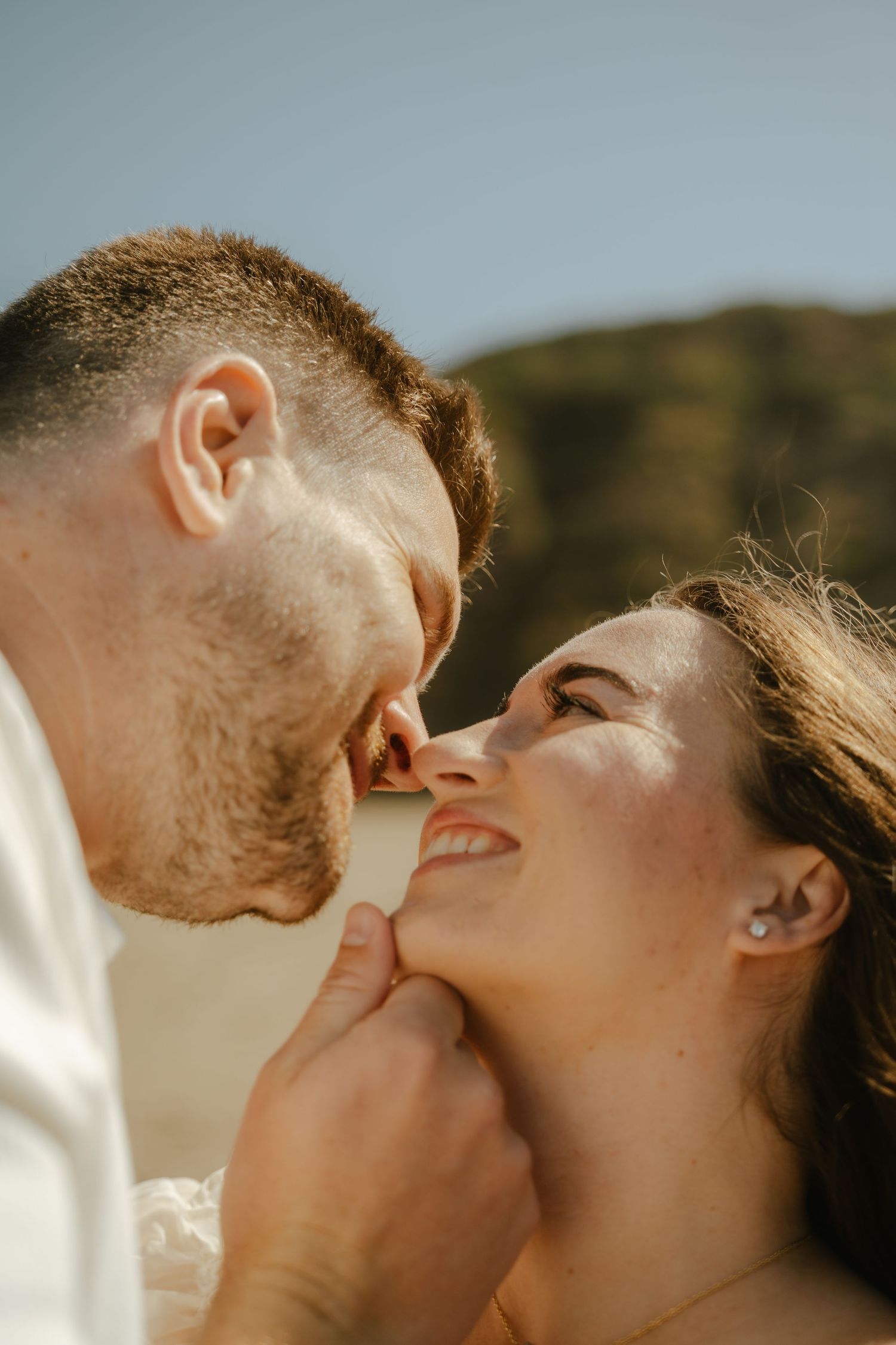 Romantic couple almost kissing outdoors, man tenderly holds womans chin as she gazes up smiling in warm sunlight.