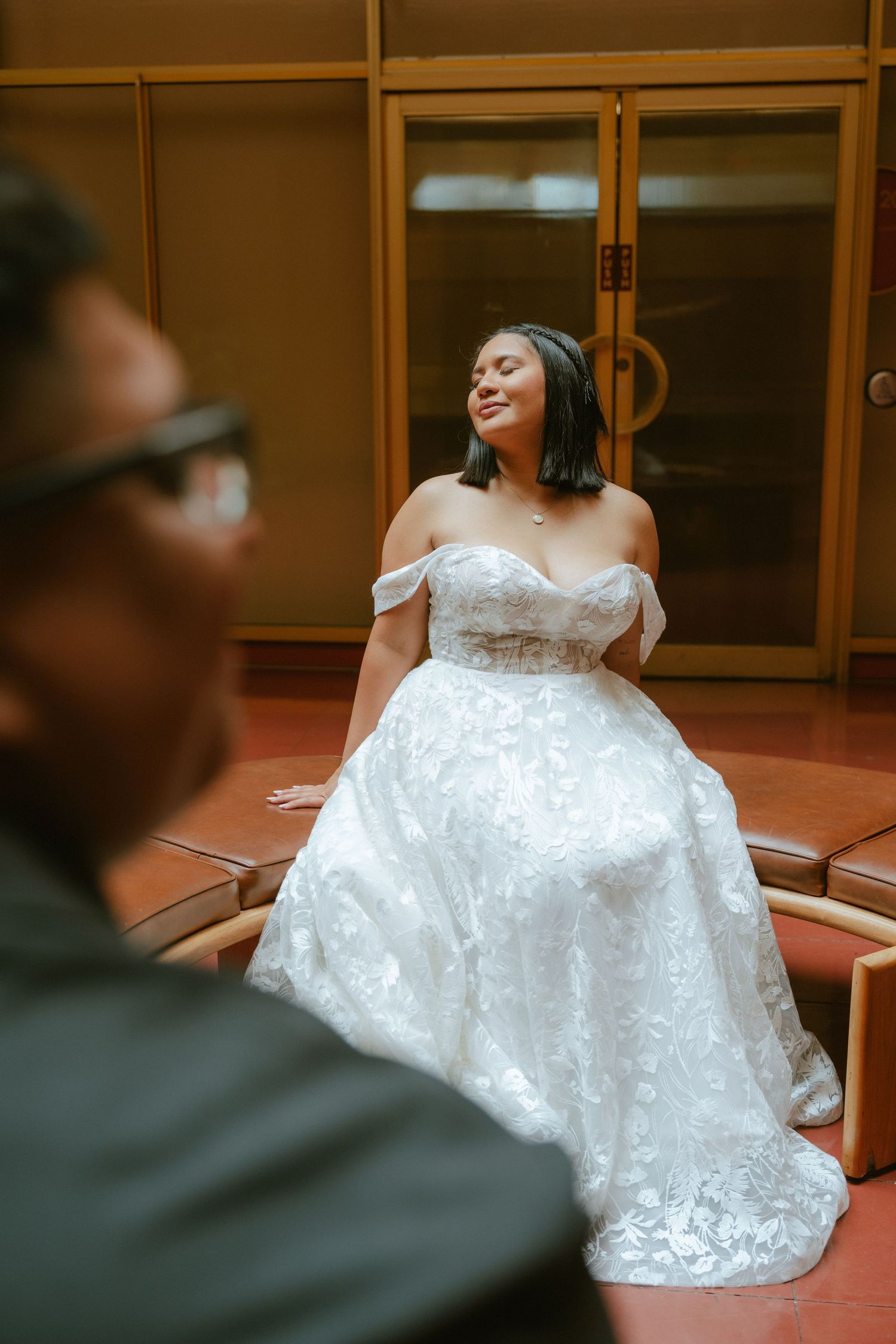 Bride in off-shoulder lace ball gown smiling joyfully while seated, groom blurred in foreground.