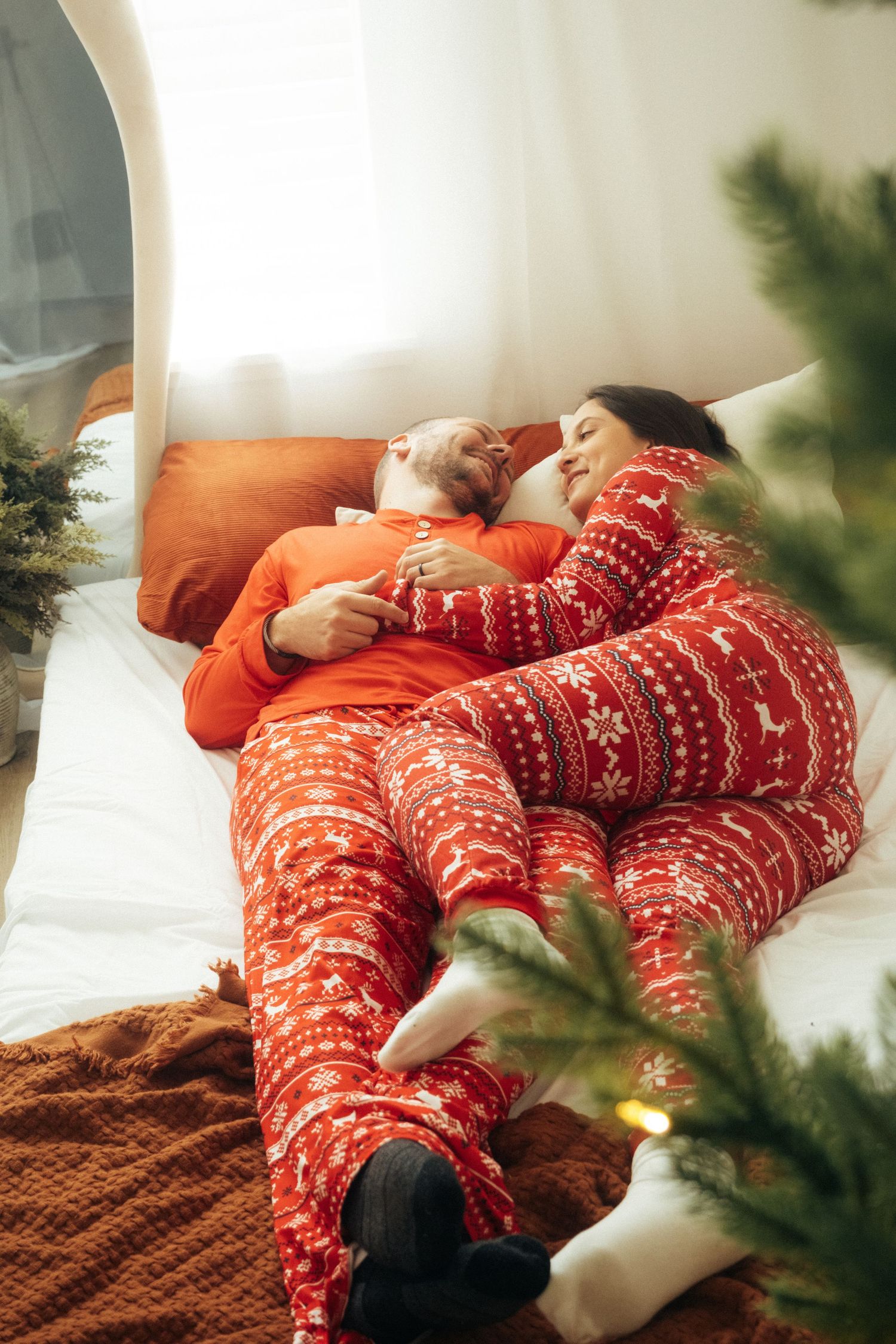 Couple in matching holiday pajamas lounging together on white mattress with Christmas decor.