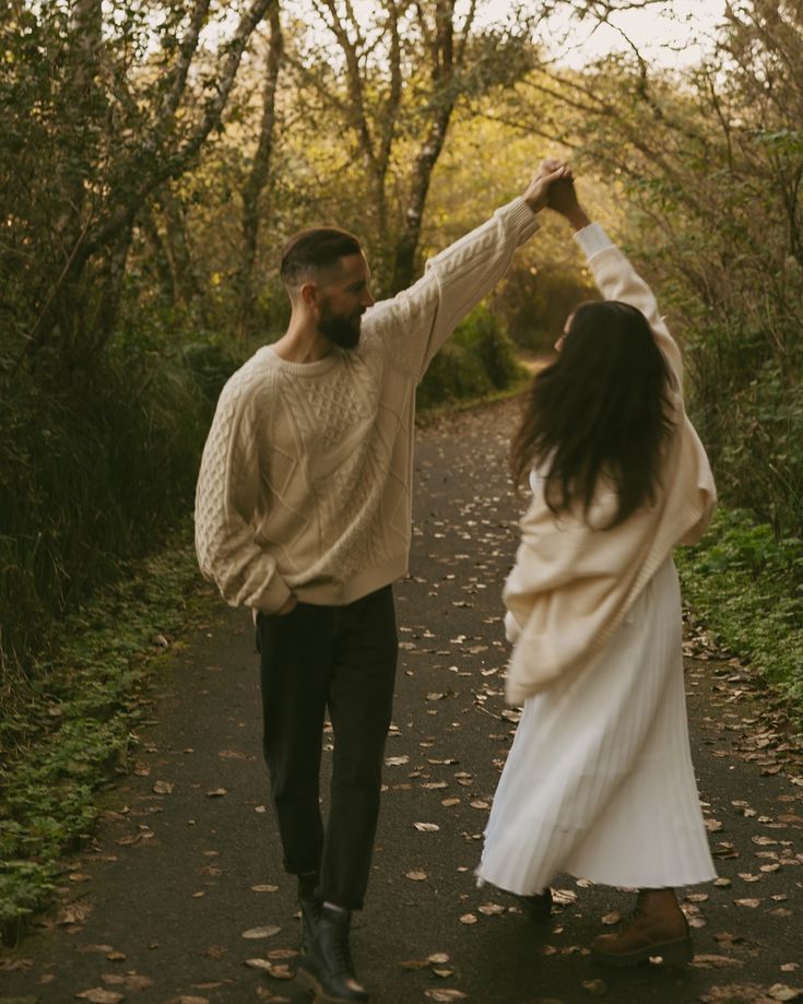 Two people dance joyfully on an autumn path surrounded by trees and fallen leaves.