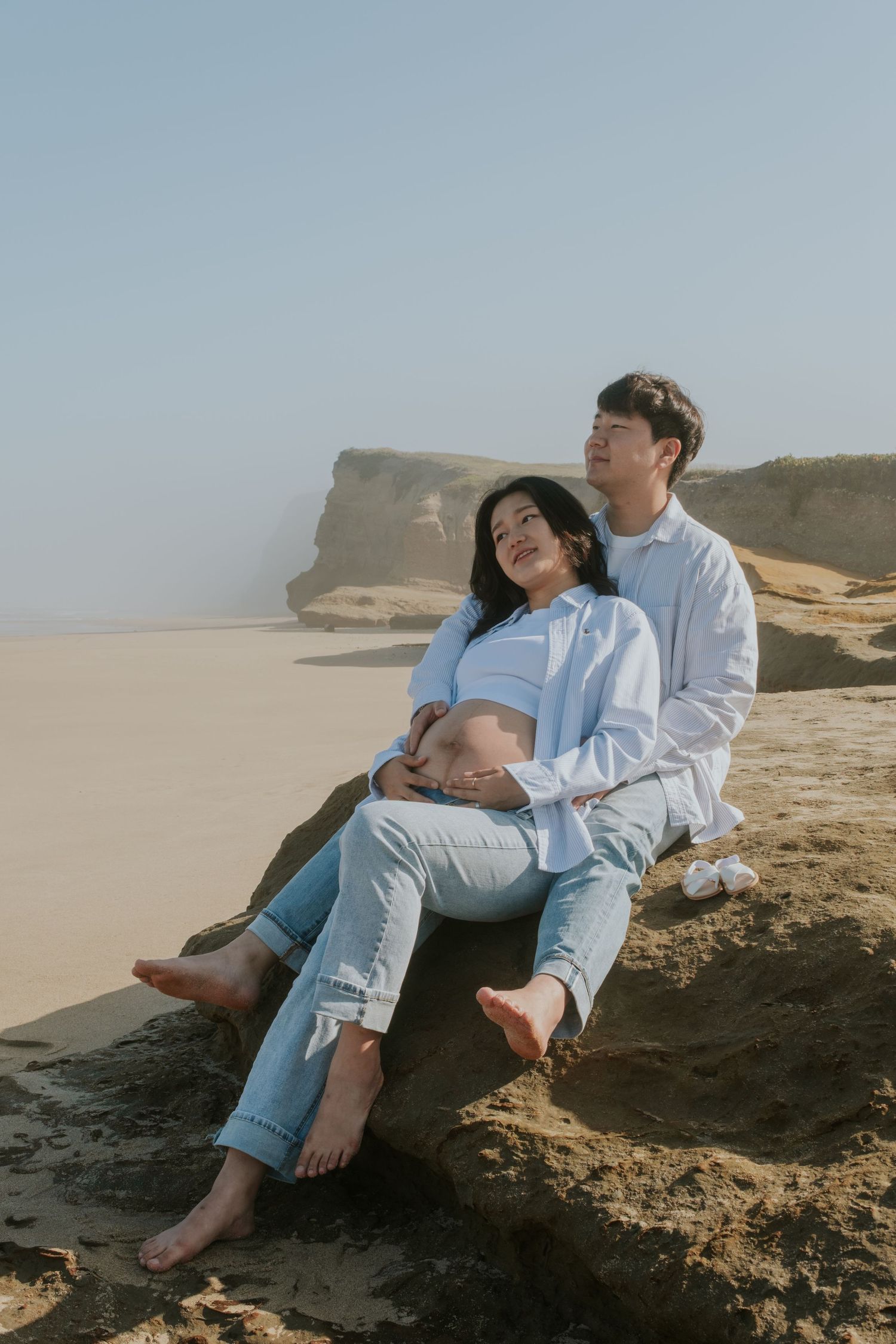 A couple relaxes on coastal rocks overlooking a sandy beach during a sunset photoshoot.