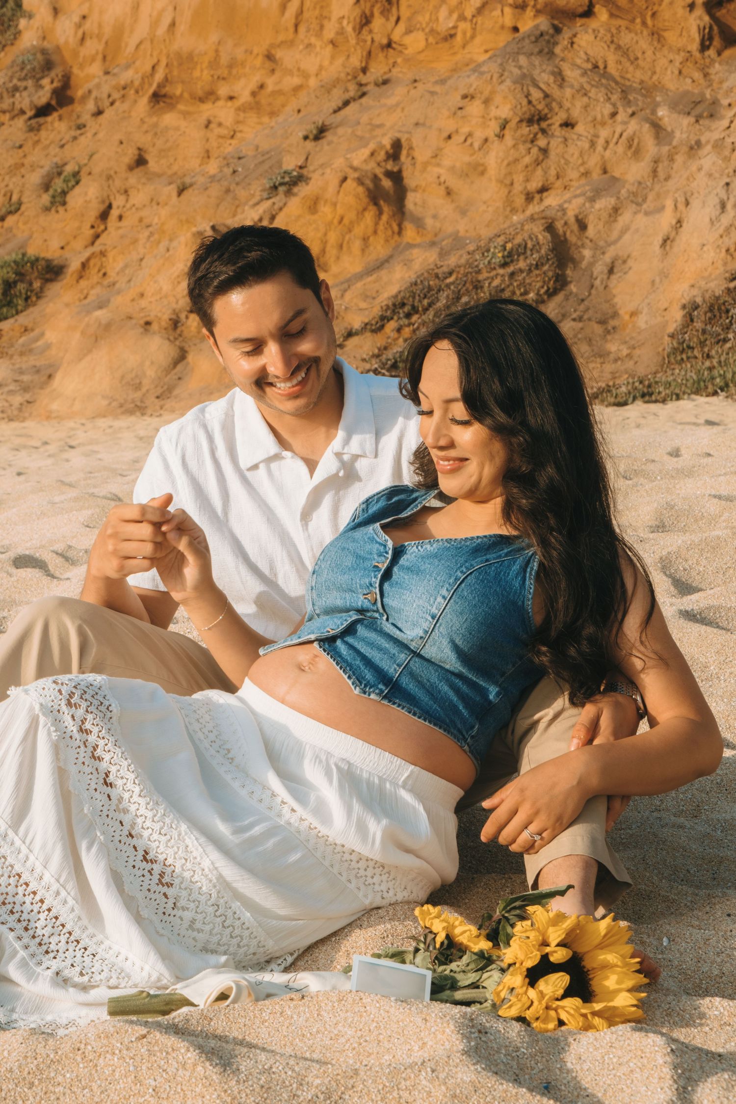 A couple relaxes on the beach with sunflowers beside them against golden cliffs.