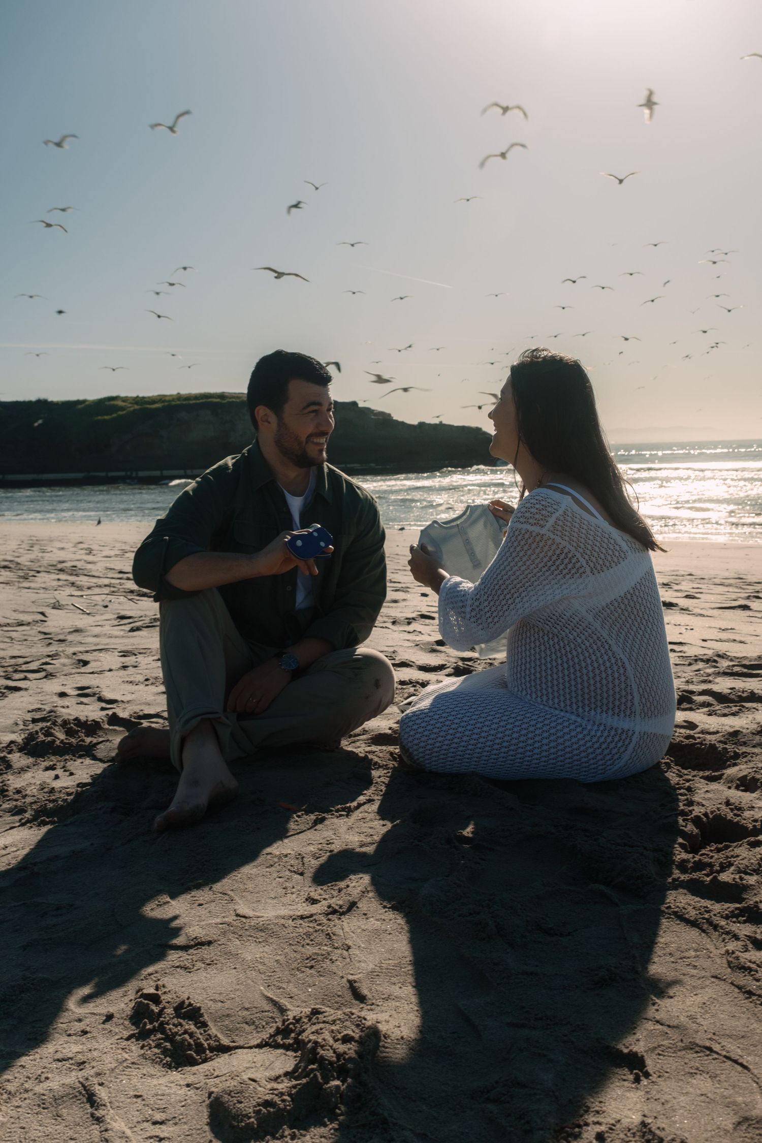 A couple sits on the beach at sunset with seagulls flying overhead.
