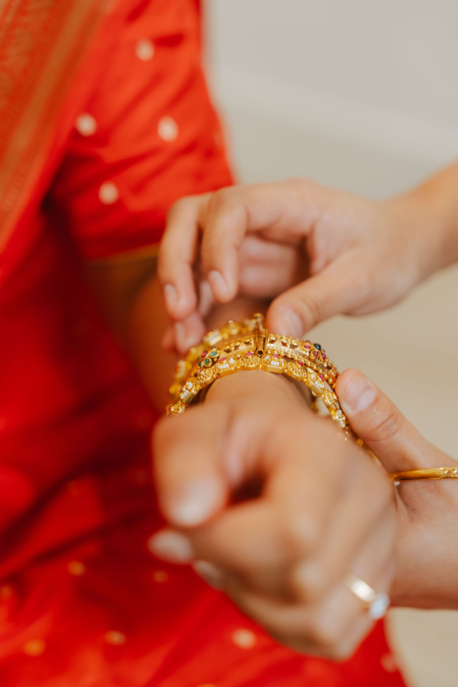 Gold bangles being placed on a bride's wrist wearing a red saree at an Indian wedding ceremony.