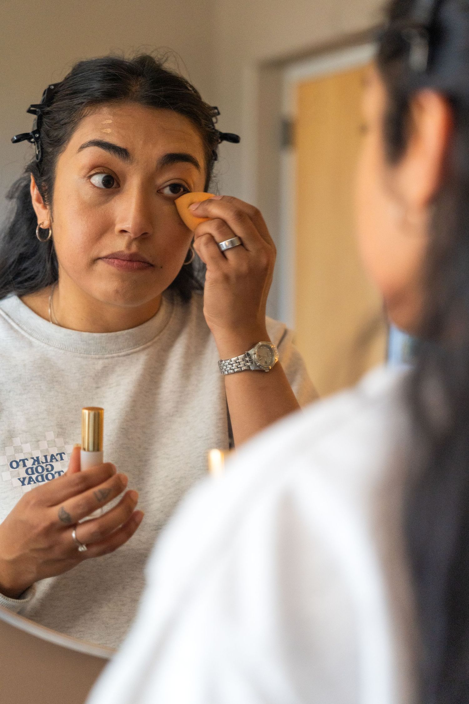 Woman applying foundation with a sponge while holding a lipstick tube, getting ready in front of a mirror.