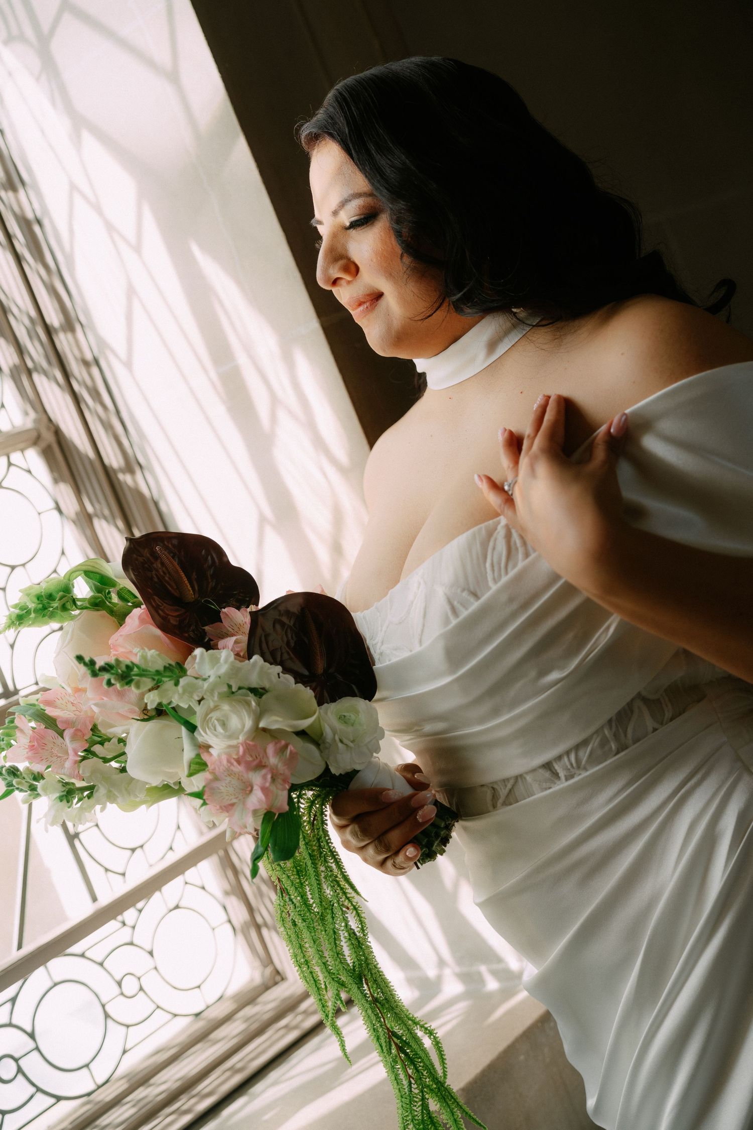 Bride in white strapless gown holding cascading floral bouquet with pink and dark flowers by ornate window.