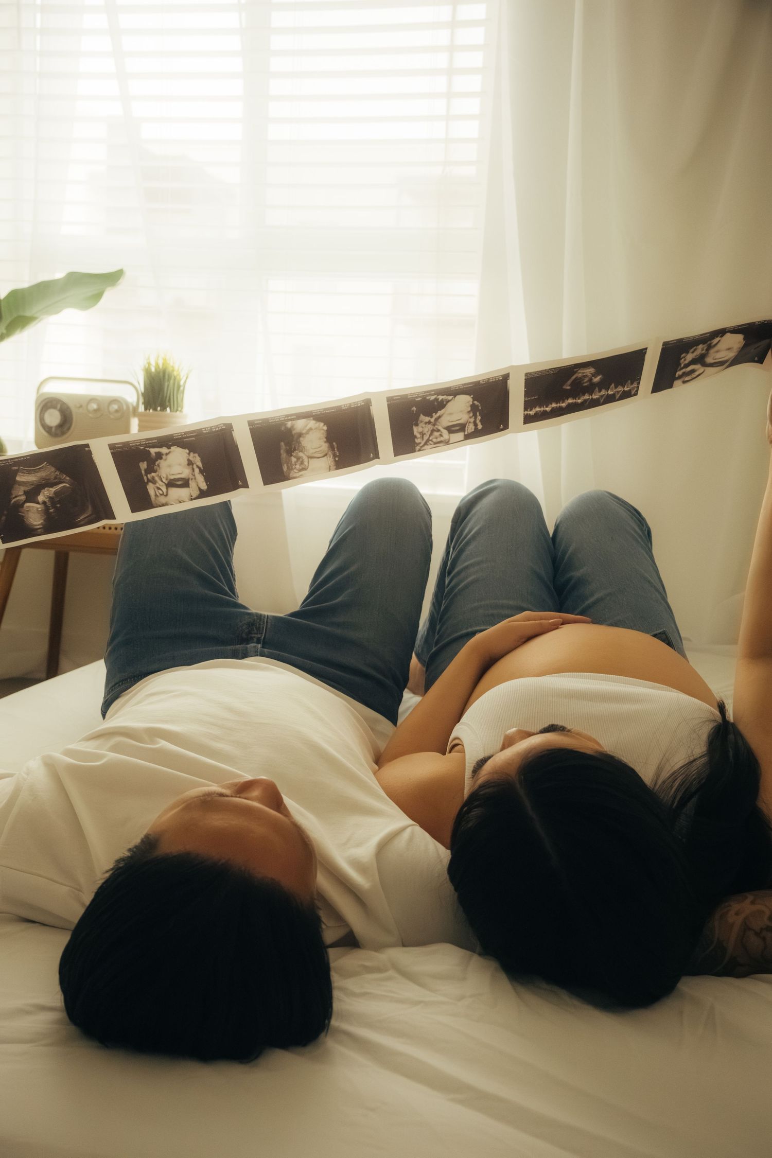 Couple lying on bed with ultrasound photo strip displayed above them near window.