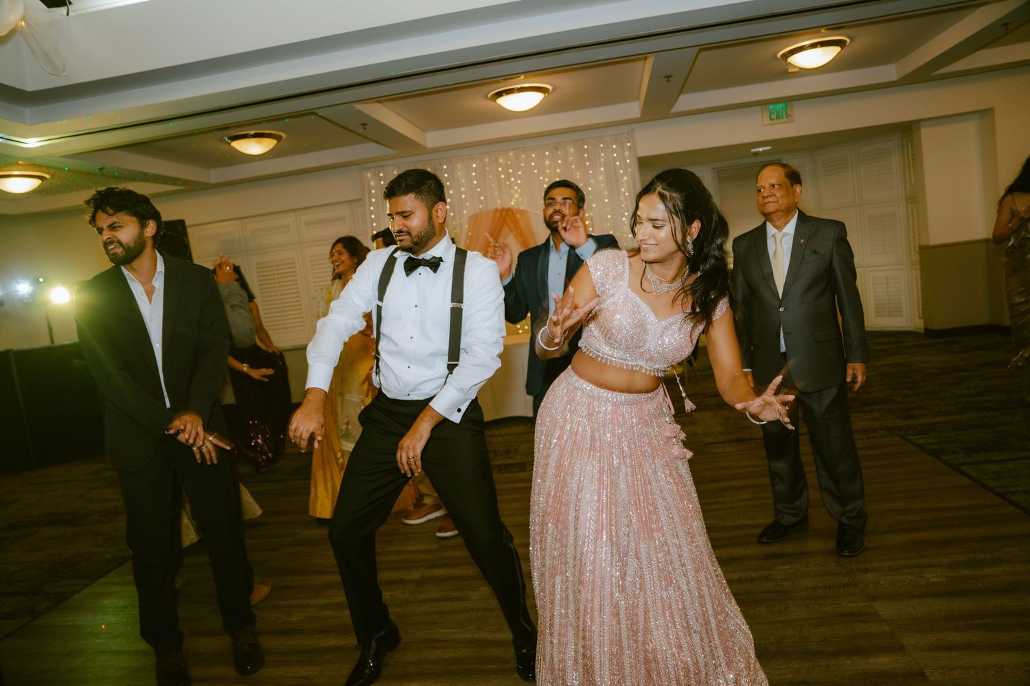 Bride and groom dancing joyfully at Indian wedding reception with guests in elegant attire in ballroom.