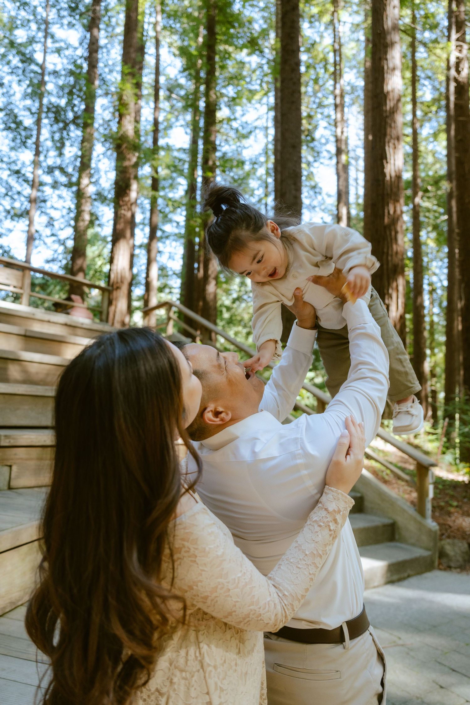 A joyful family moment in a redwood forest, with a man lifting a laughing toddler while a woman embraces him.