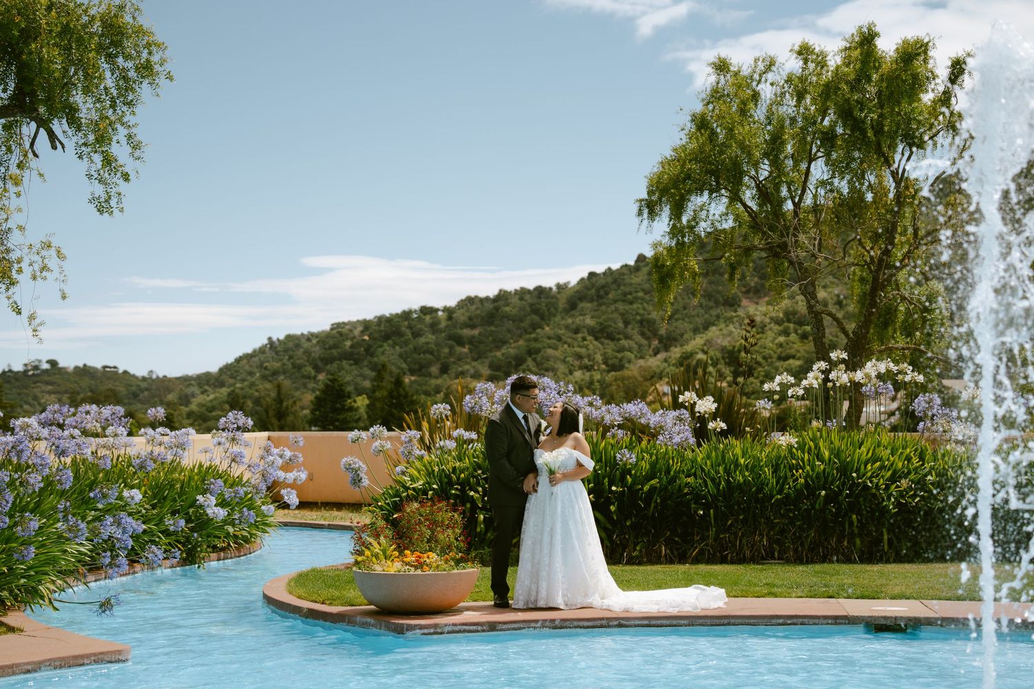 Bride and groom share romantic moment on island surrounded by fountain with purple flowers and hills beyond.