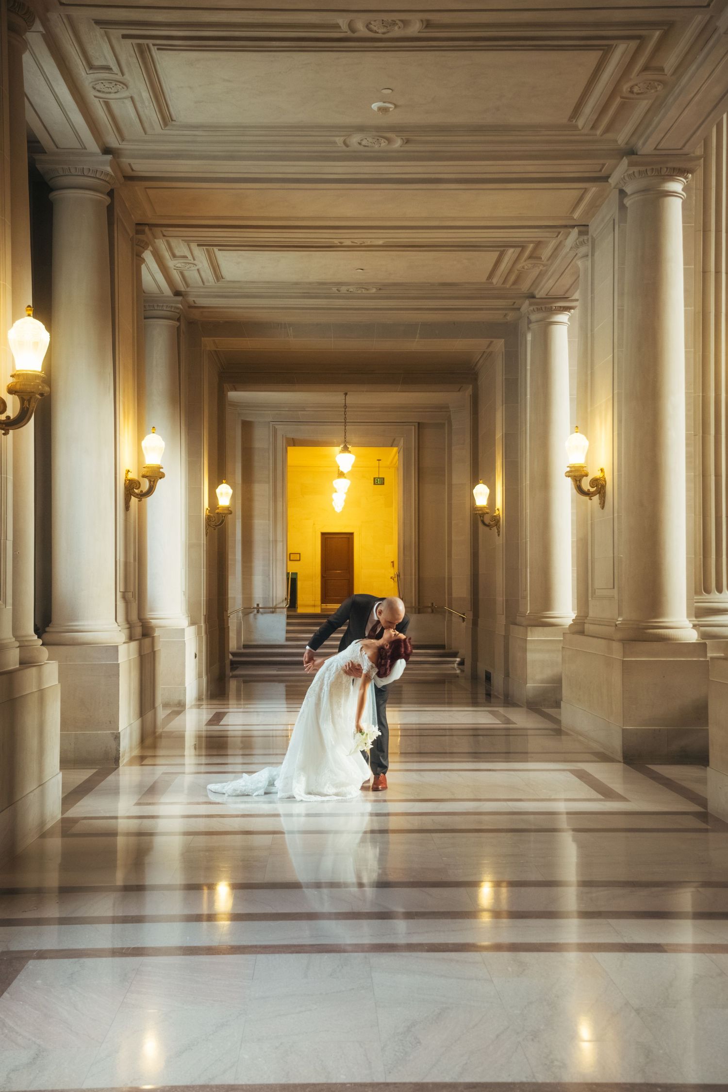 Elegant wedding couple walking through grand hallway with ornate columns and warm lighting.
