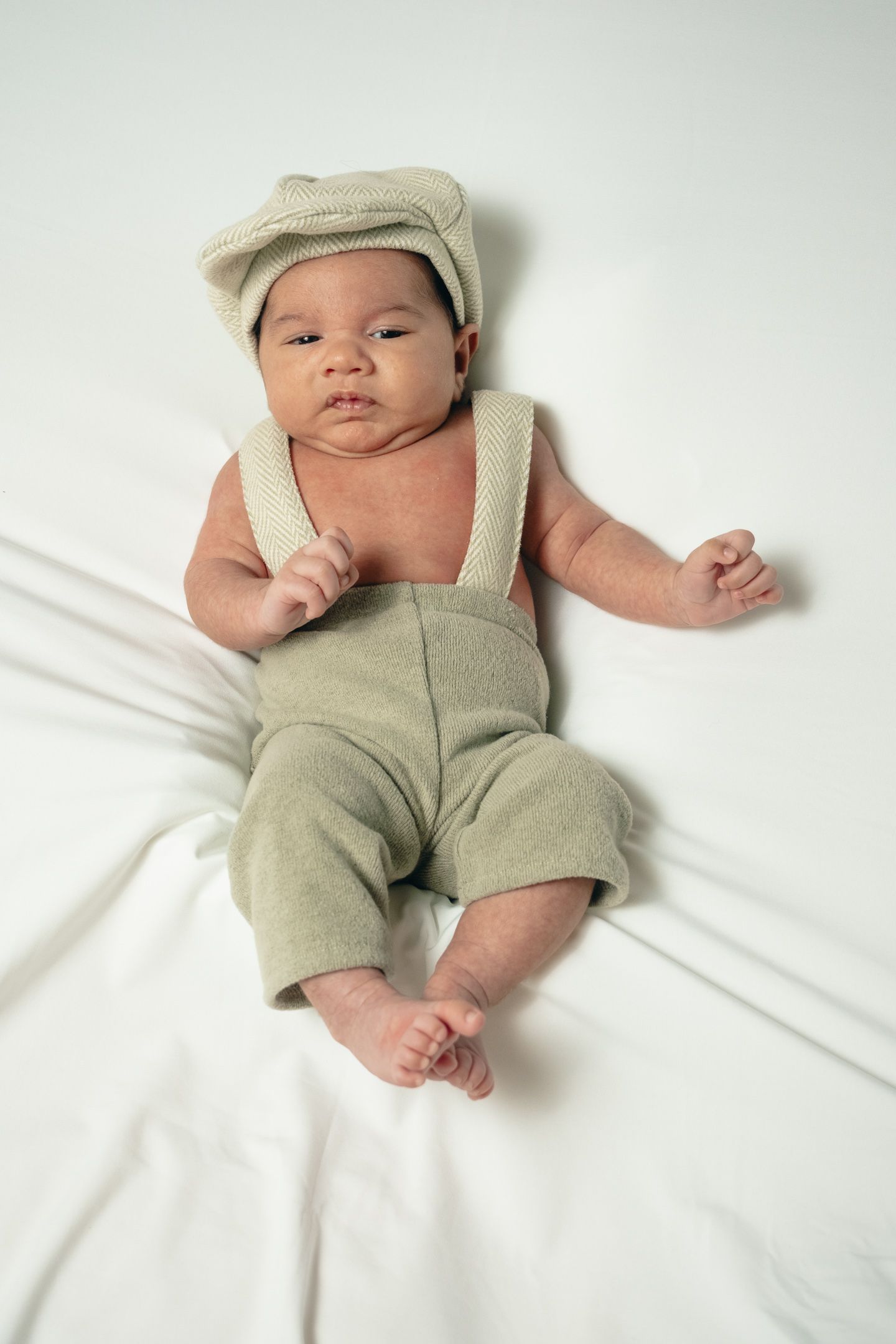 Baby in beige overalls and cap lying on white background.