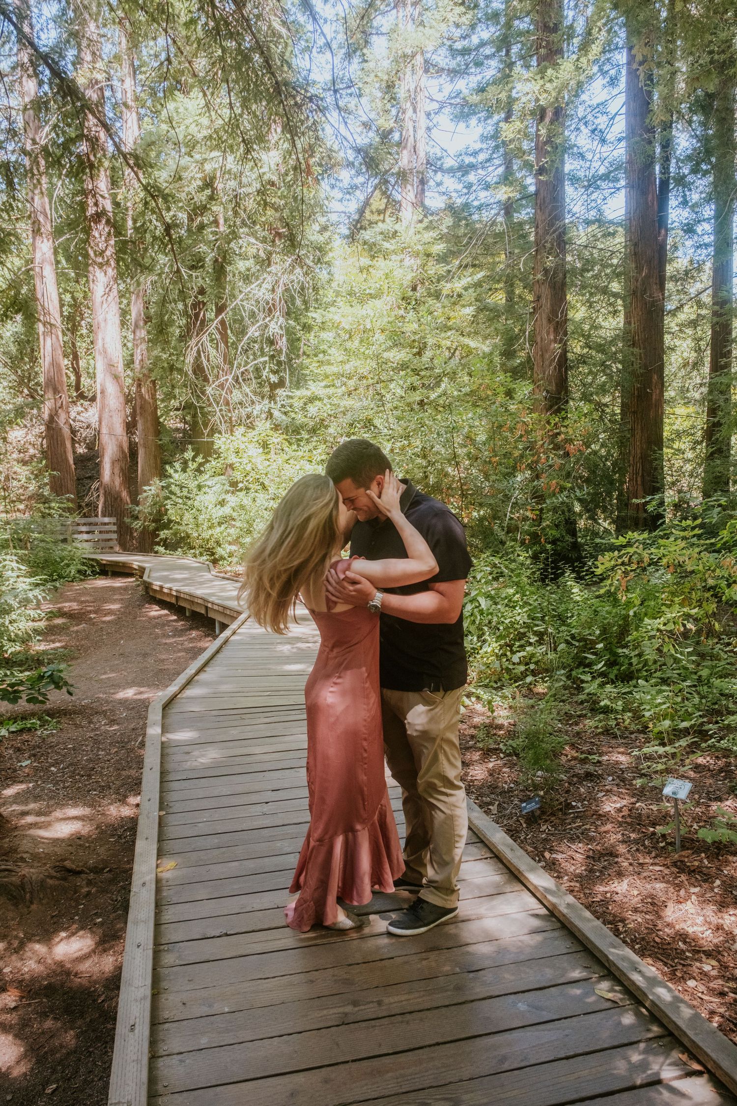 A couple dances romantically on a wooden boardwalk surrounded by towering trees.
