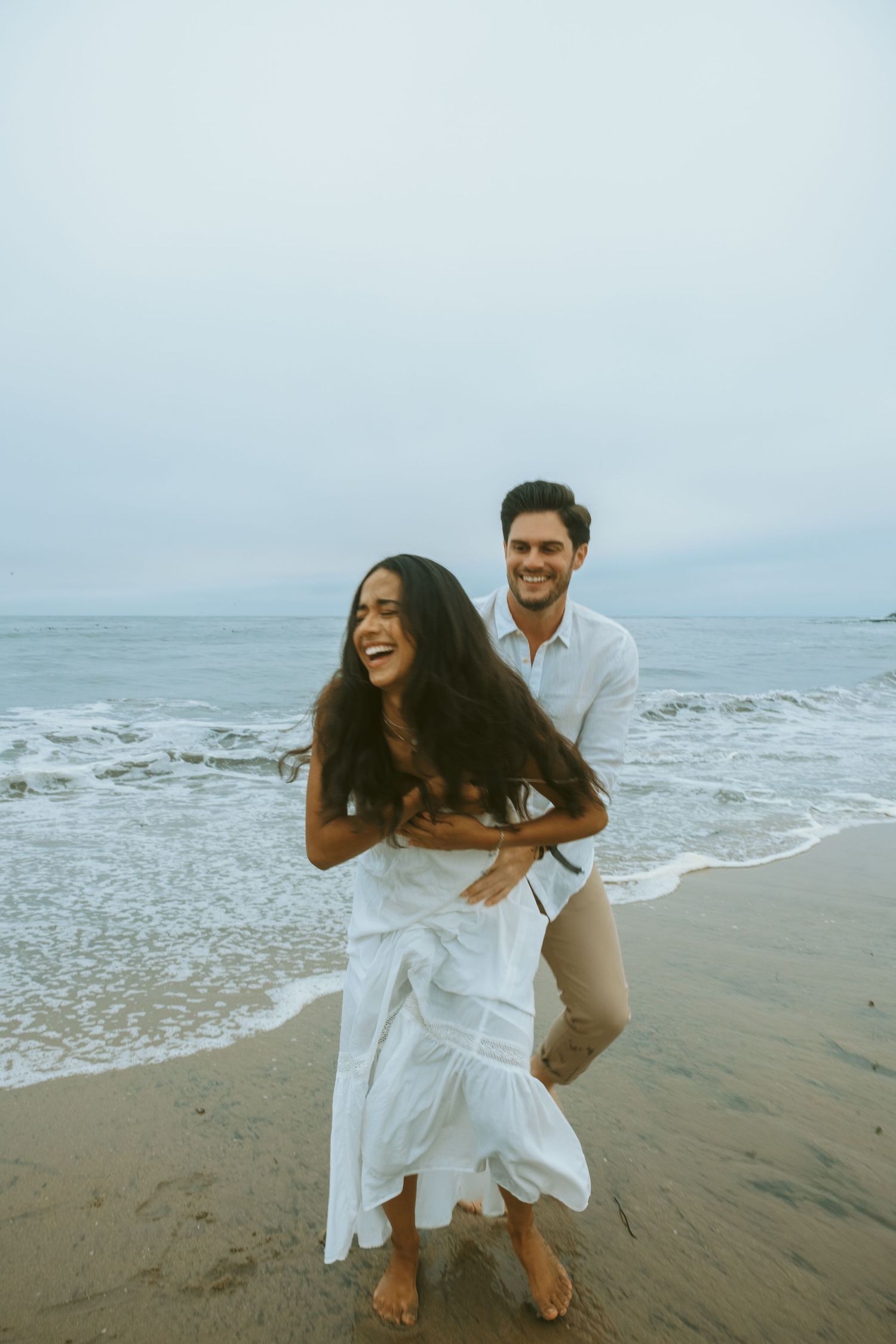 A playful couple in white outfits frolicking on a beach with waves in the background.