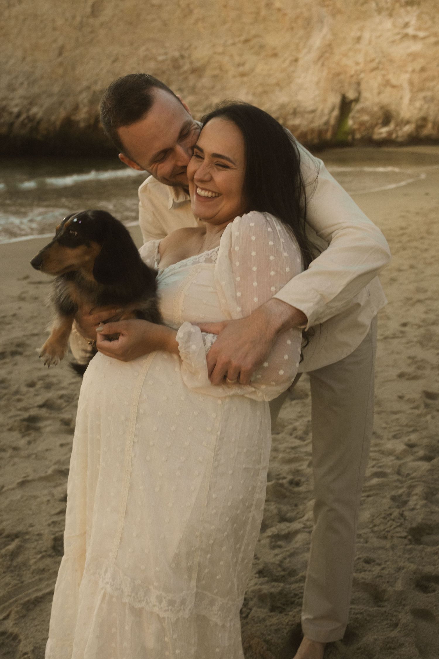 Couple embracing on a beach with rocky cliffs in the background.