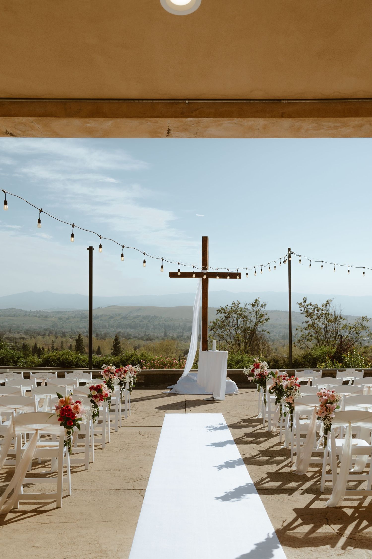 Elegant outdoor wedding aisle with white chairs, floral arrangements, wooden cross, and mountain backdrop.