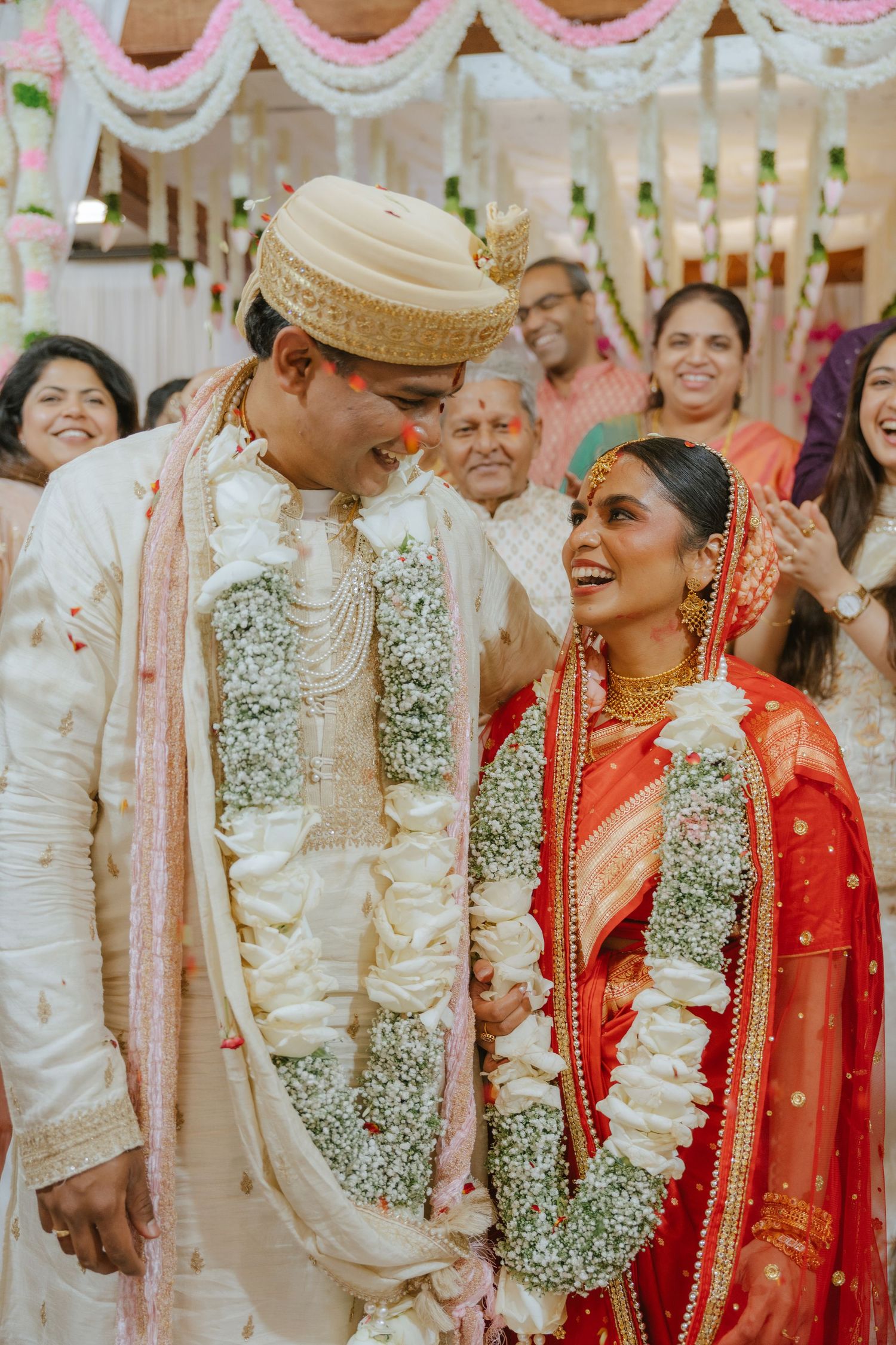 Joyful Indian bride in red saree and groom in white sherwani exchange garlands at traditional Hindu wedding ceremony.