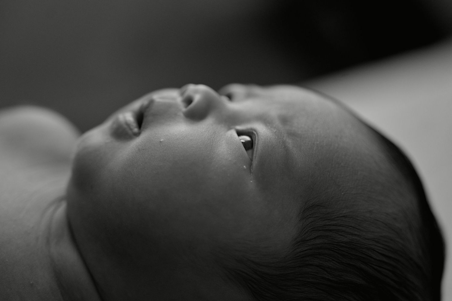 Black and white close-up portrait of a newborn baby lying down, eyes open, soft skin detail.