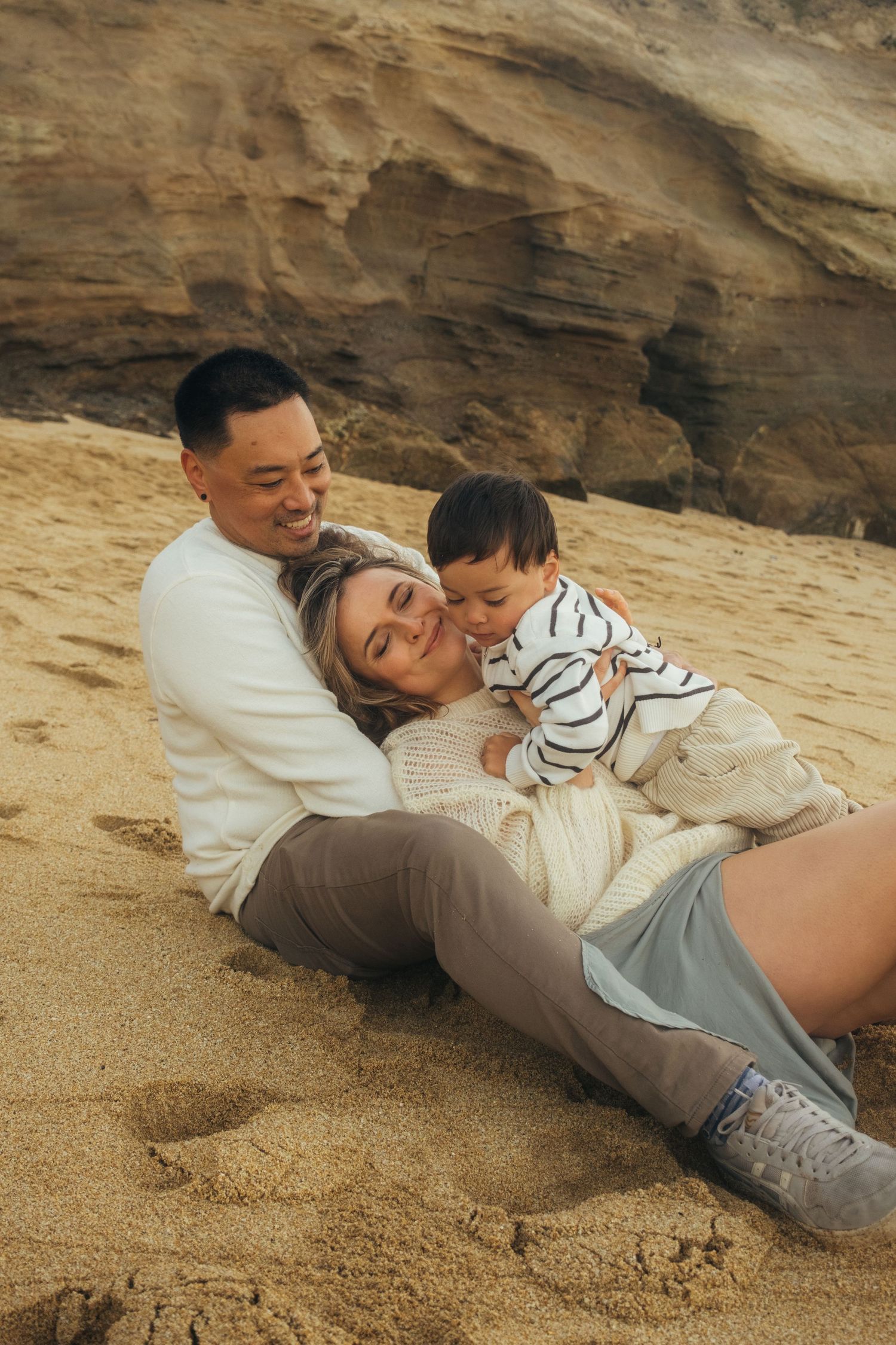 Family of three sitting on sandy beach, parents smiling as toddler laughs, rocky cliffs in background.