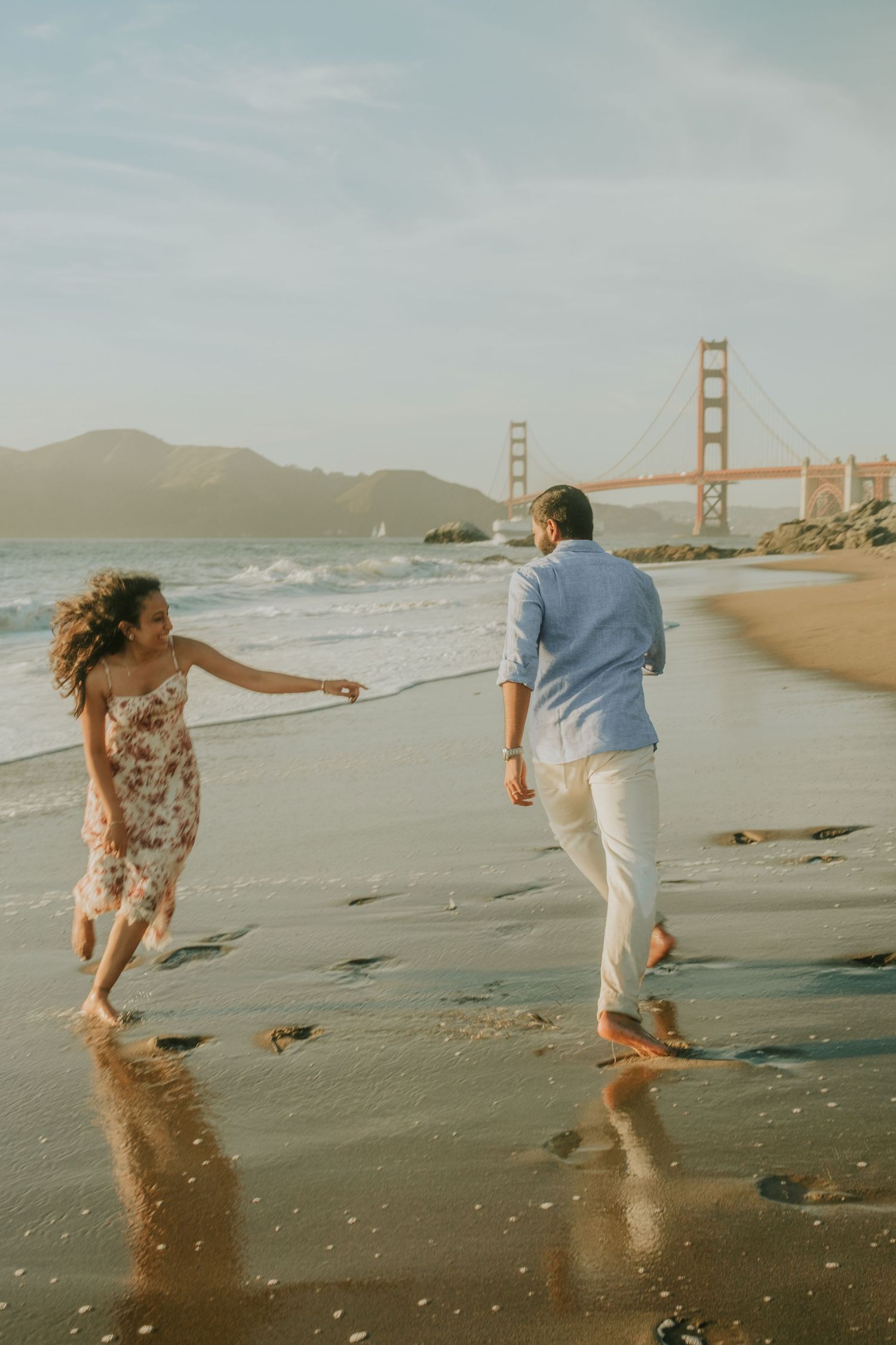 Couple holding hands walking barefoot on Baker Beach with the Golden Gate Bridge in the background.